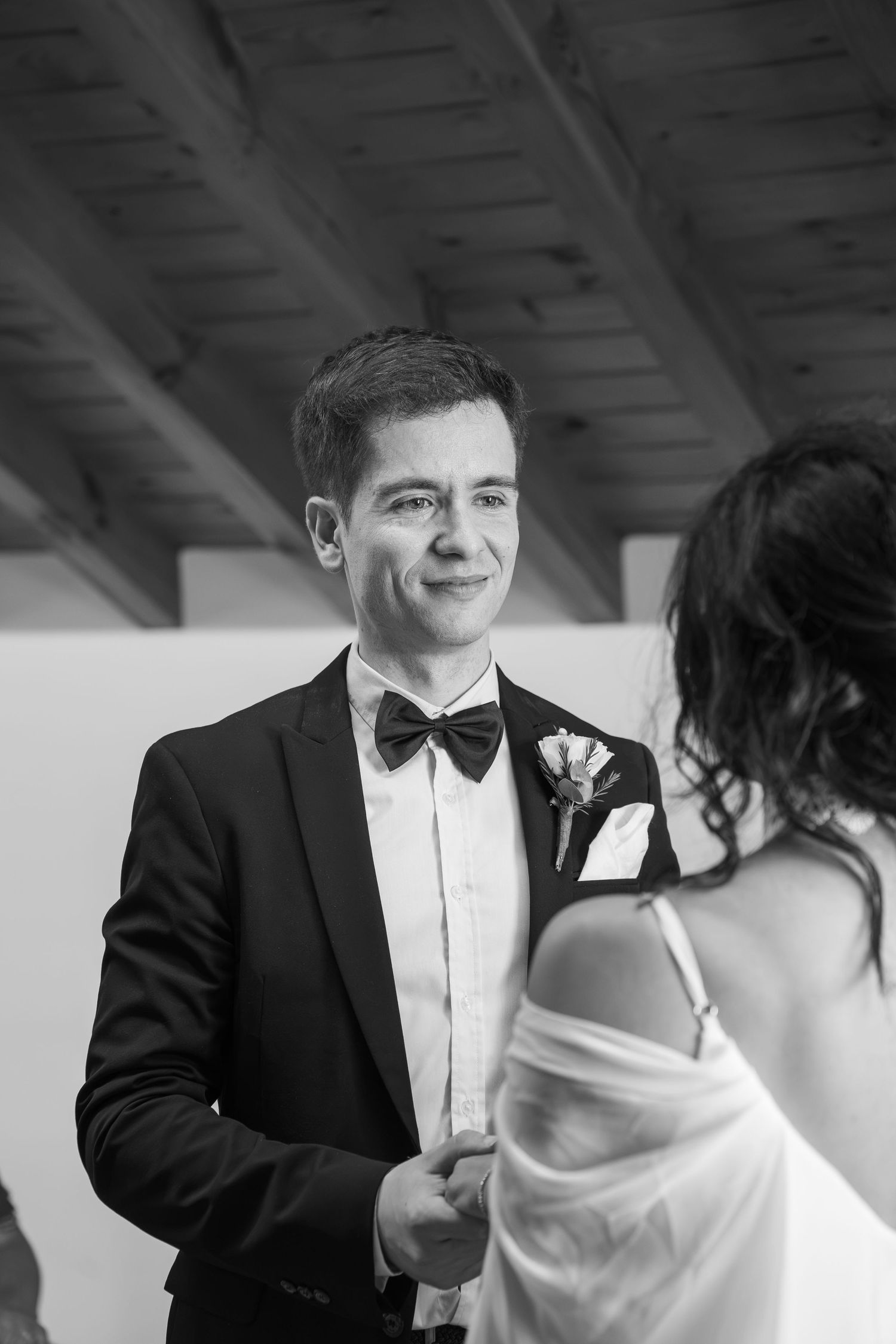 Smiling groom in a black tuxedo with bow tie and boutonniere holds hands with his bride during a wedding ceremony, in black and white.