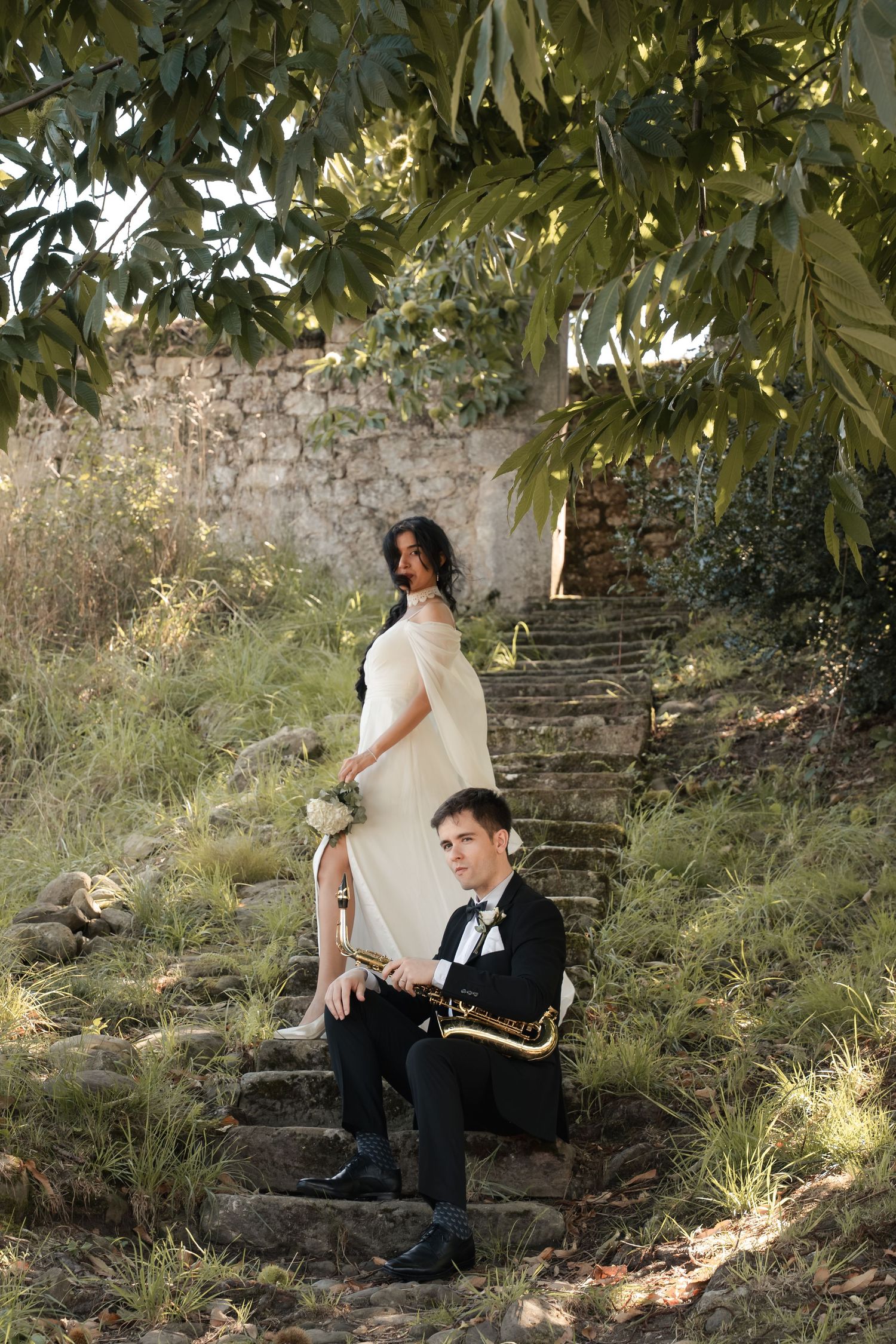 Bride in a white gown with a bouquet stands on stone steps behind a groom in a tuxedo seated with a saxophone, framed by leafy trees.