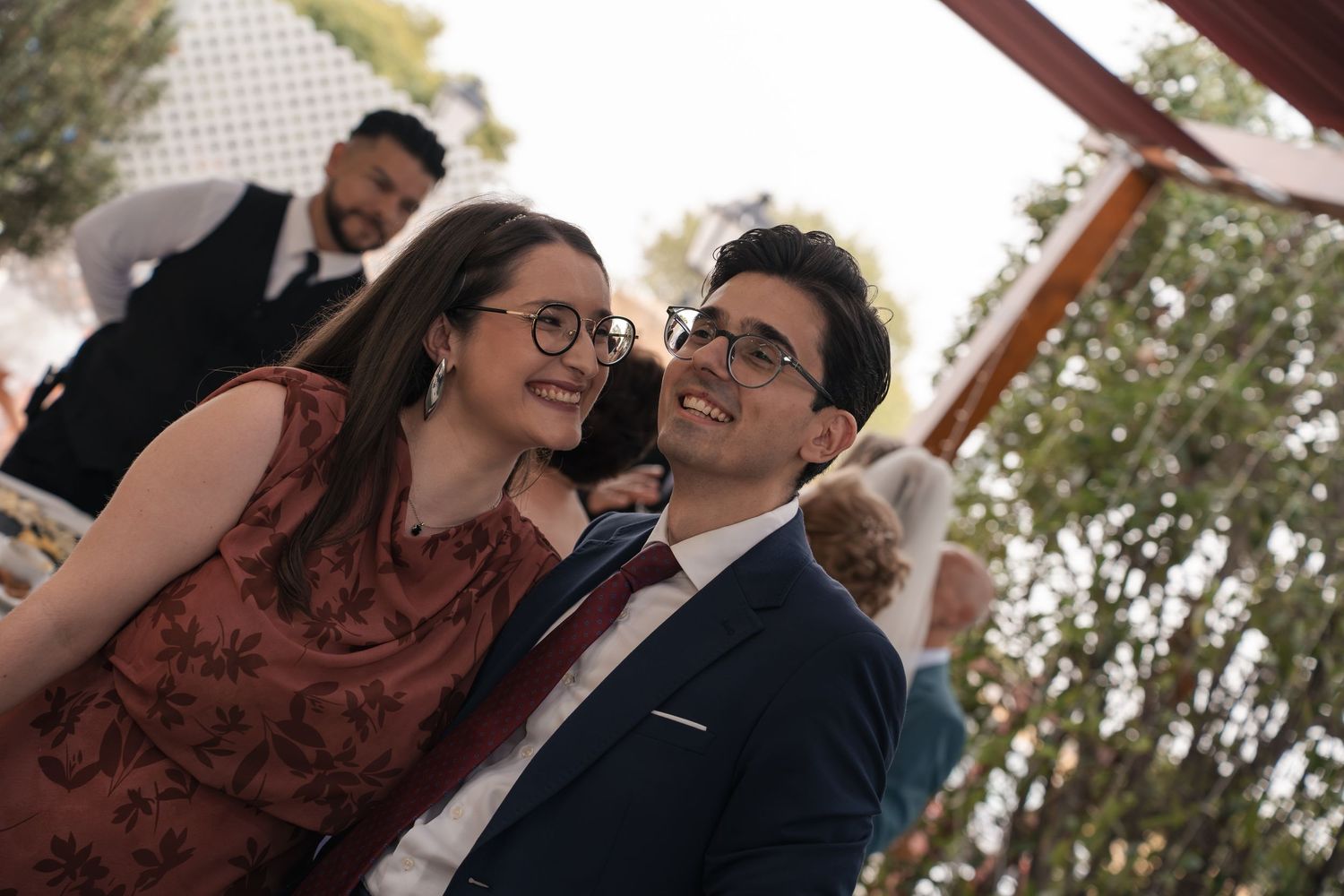 Smiling young woman in a floral dress and glasses poses beside a man in a navy suit at an outdoor celebration.