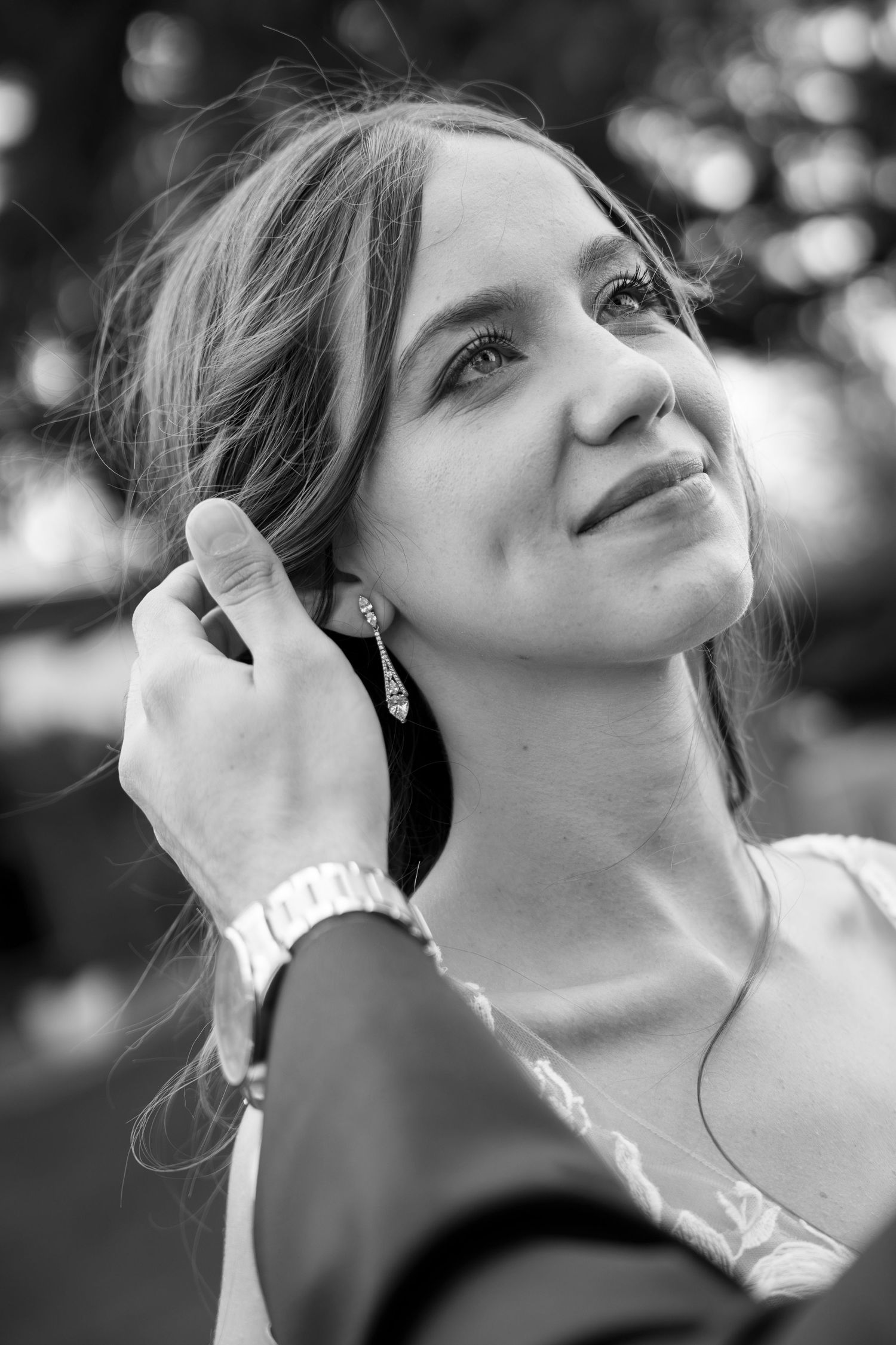 Black and white portrait of a smiling bride with drop earrings as a hand in a suit sleeve gently tucks her hair behind her ear.