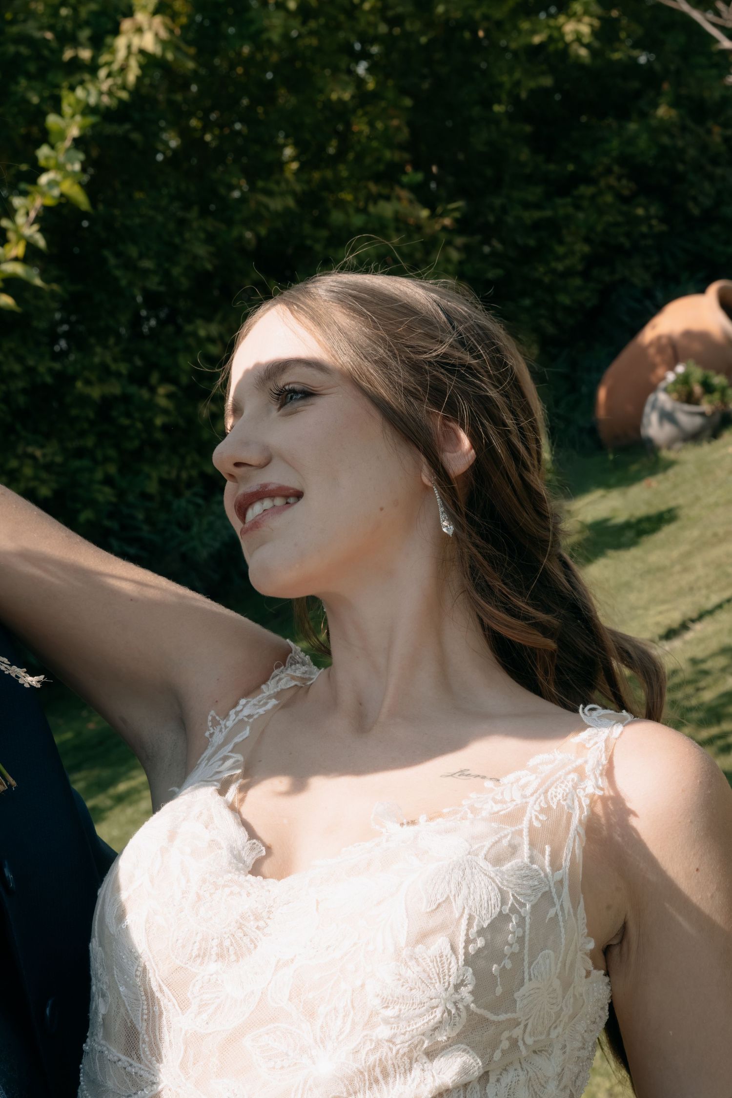 Smiling bride in a lace wedding gown with drop earrings and wavy hair, arm raised, posing outdoors in a sunlit garden.