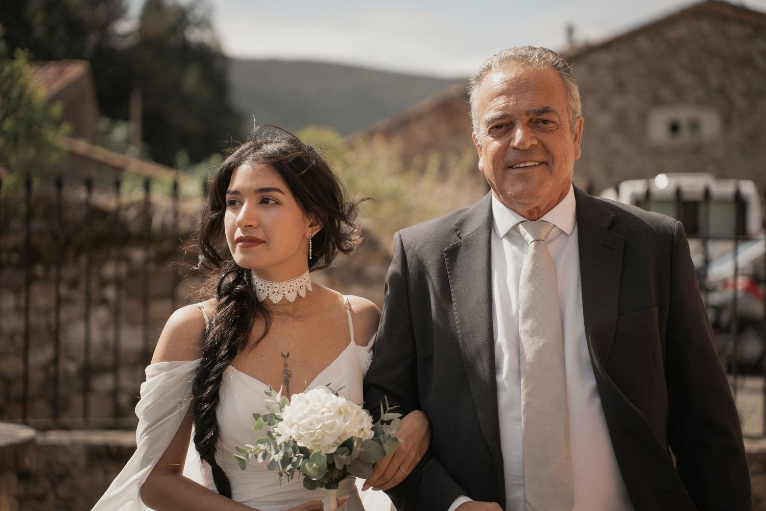 A bride in a white off-the-shoulder gown holding a white bouquet walks arm in arm with a smiling older man in a dark suit outdoors.