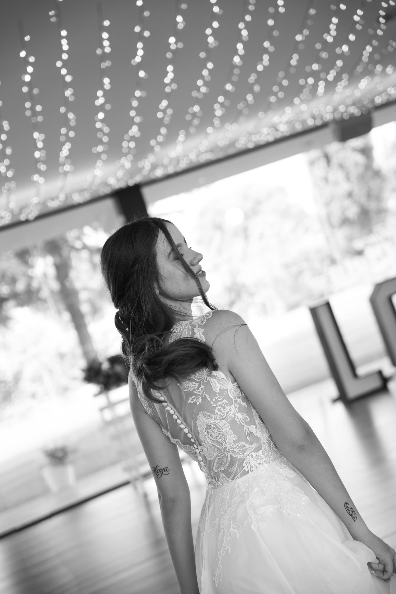 Black and white photo of a bride in a lace wedding dress with an open back, looking over her shoulder under string lights.