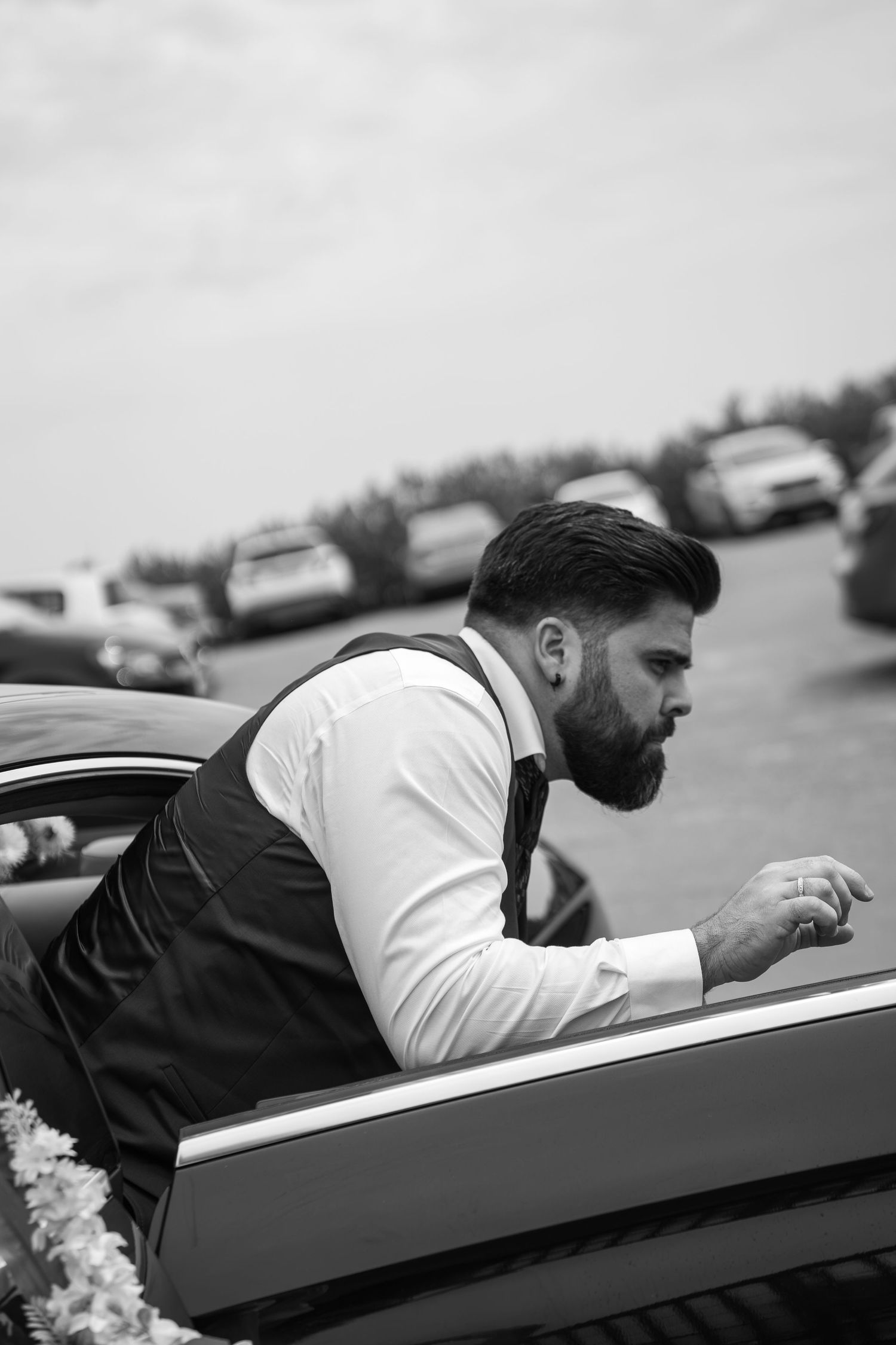 Black and white photo of a bearded man in suspenders leaning against a decorated car in a parking lot.