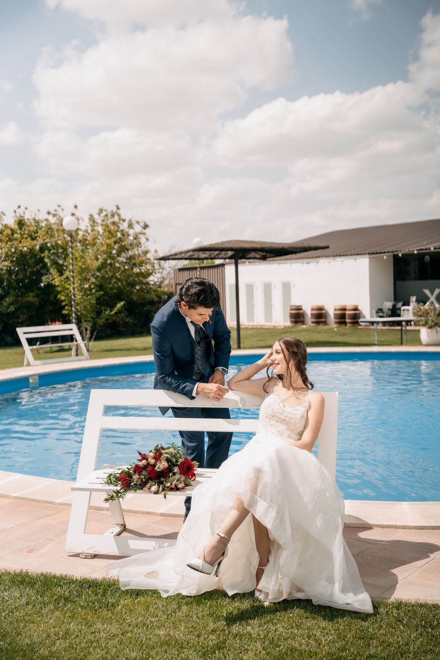 Bride in a lace wedding gown sits on a white bench beside a pool with a red bouquet as the groom in a navy suit leans toward her.