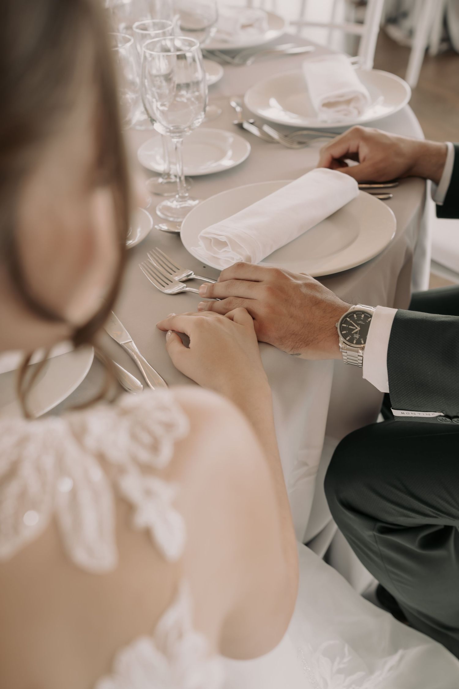 Bride in a lace wedding dress holds hands with a groom in a gray suit and wristwatch at an elegantly set reception table.
