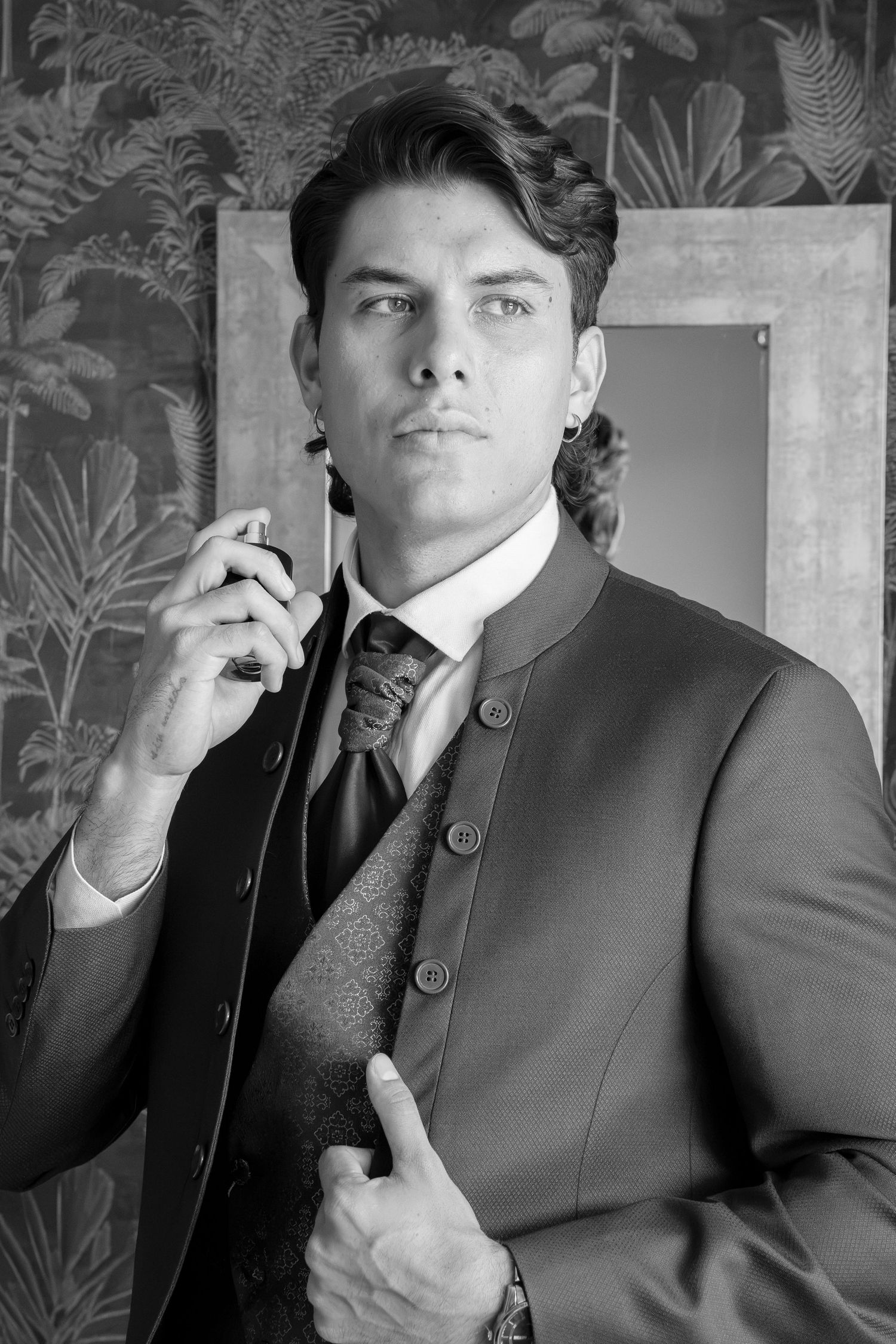 Black and white portrait of a man in a buttoned collarless jacket, patterned vest and tie, holding a perfume bottle against botanical wallpa