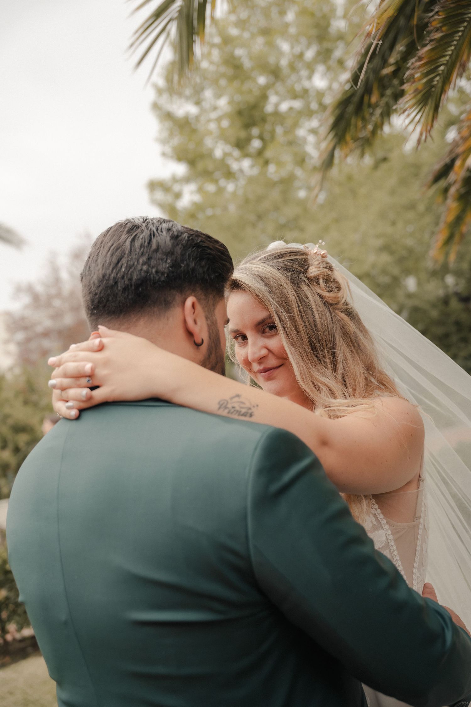 Smiling blonde bride in veil embracing groom in green suit outdoors near palm tree.
