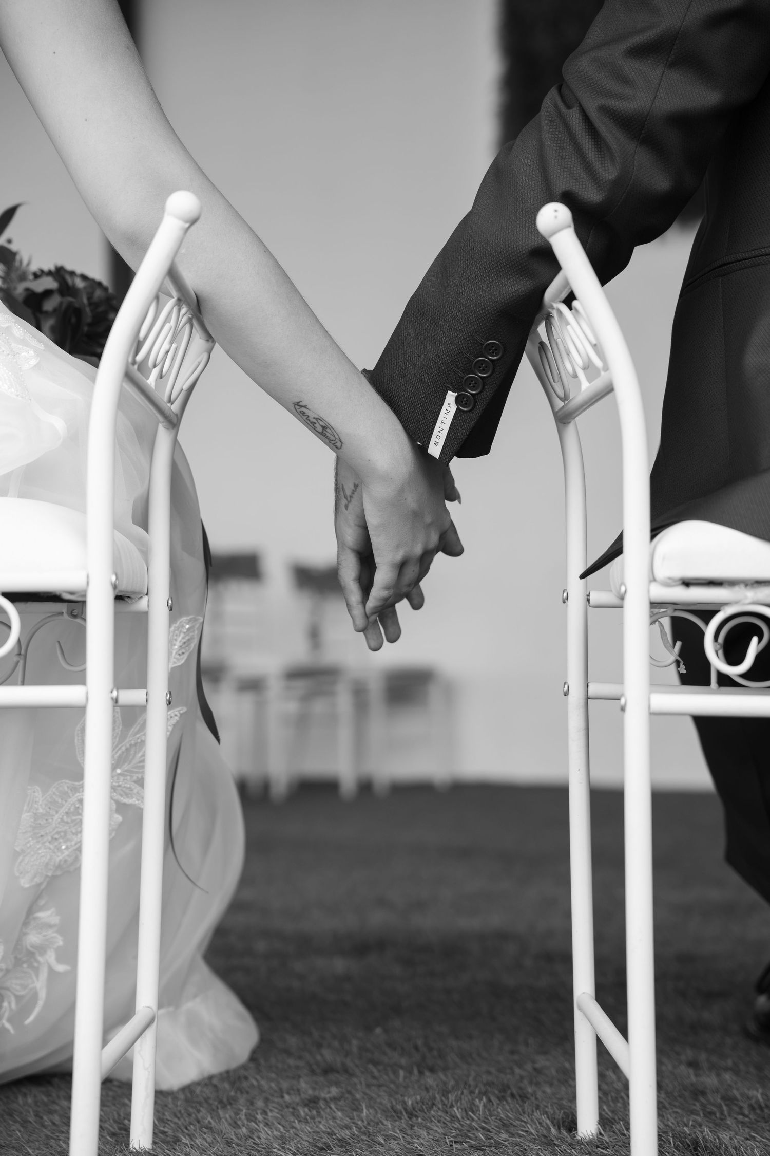 Black and white photo of a bride and groom holding hands between two white wrought iron chairs during an outdoor wedding ceremony.