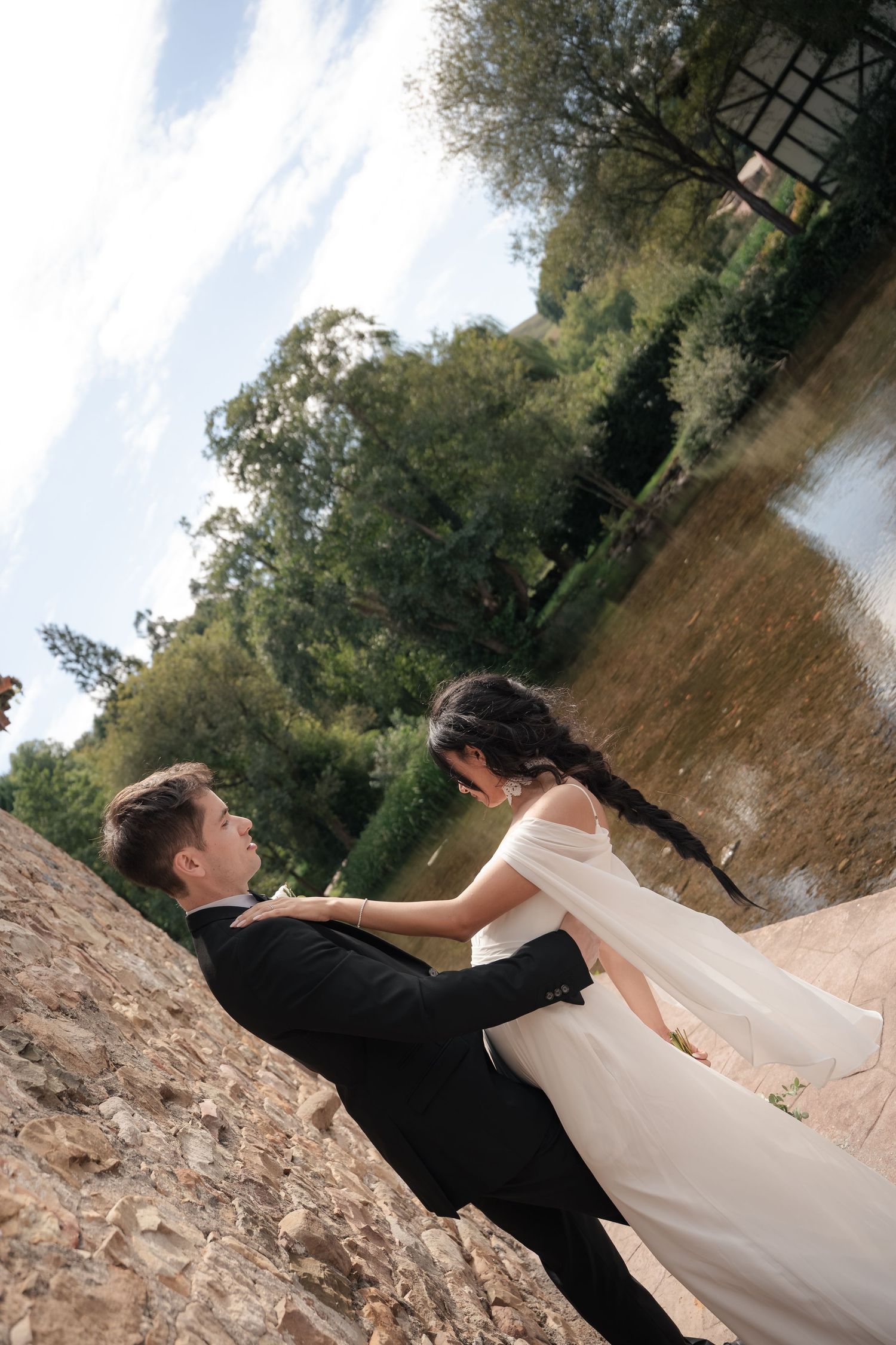 Bride in a white gown and groom in a dark suit embrace in a tilted wedding photo beside a calm pond with lush green trees.