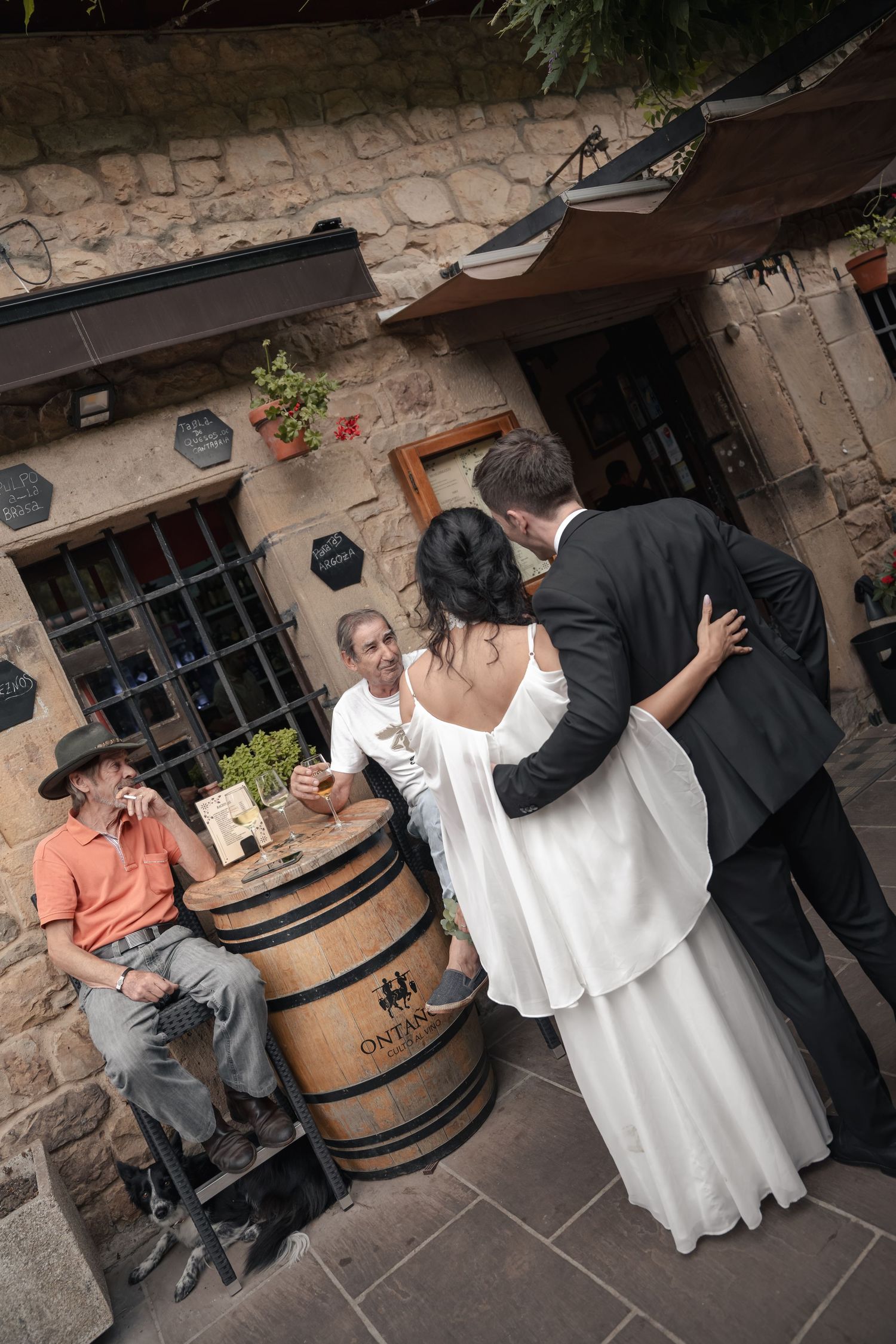 Bride in a flowing white dress embraces a groom in a dark suit beside a wine barrel, with two men chatting near a rustic stone building.