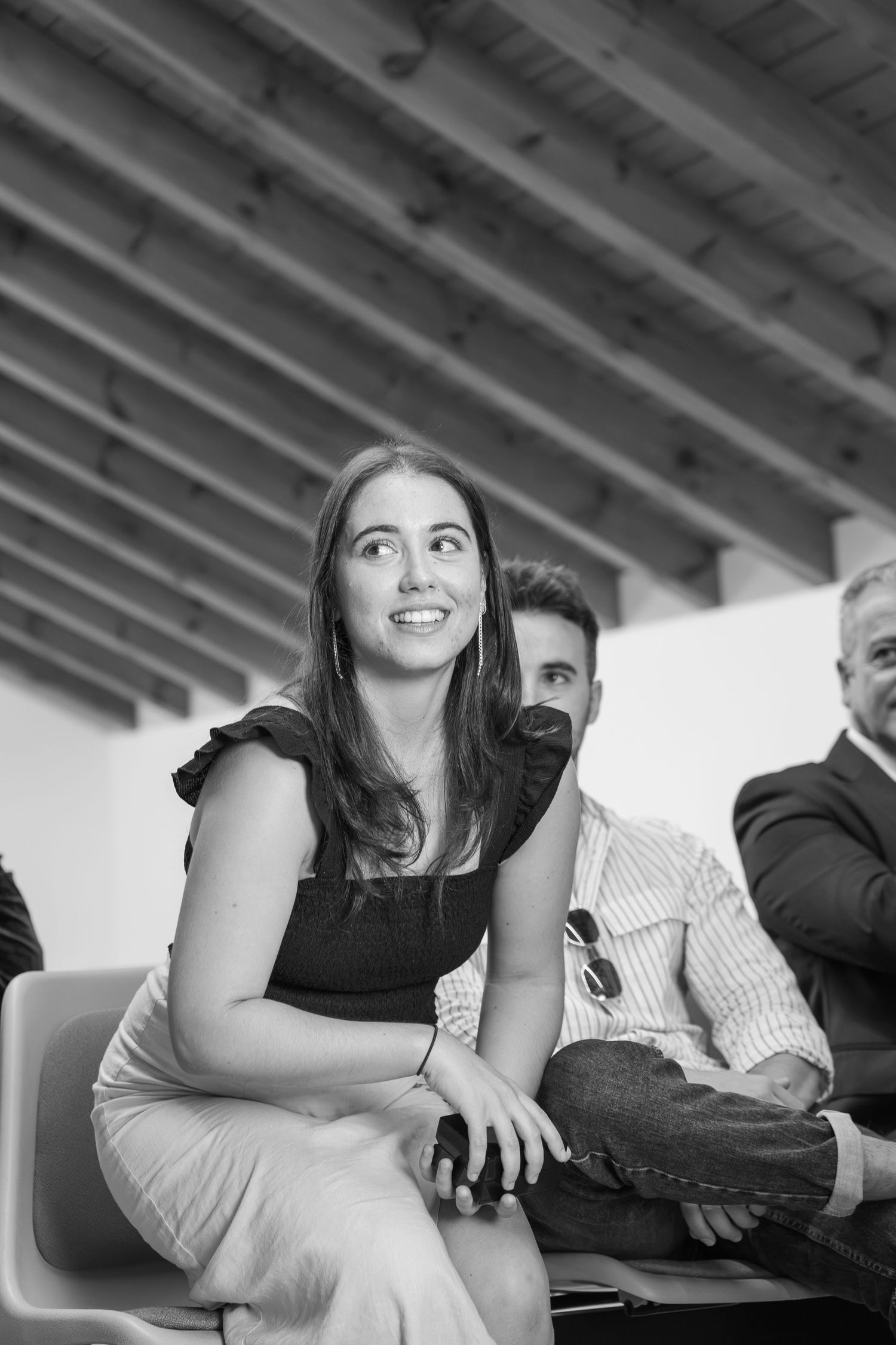 Black and white photo of a smiling young woman with long hair seated in a chair, with two men beside her under a wooden beamed ceiling.