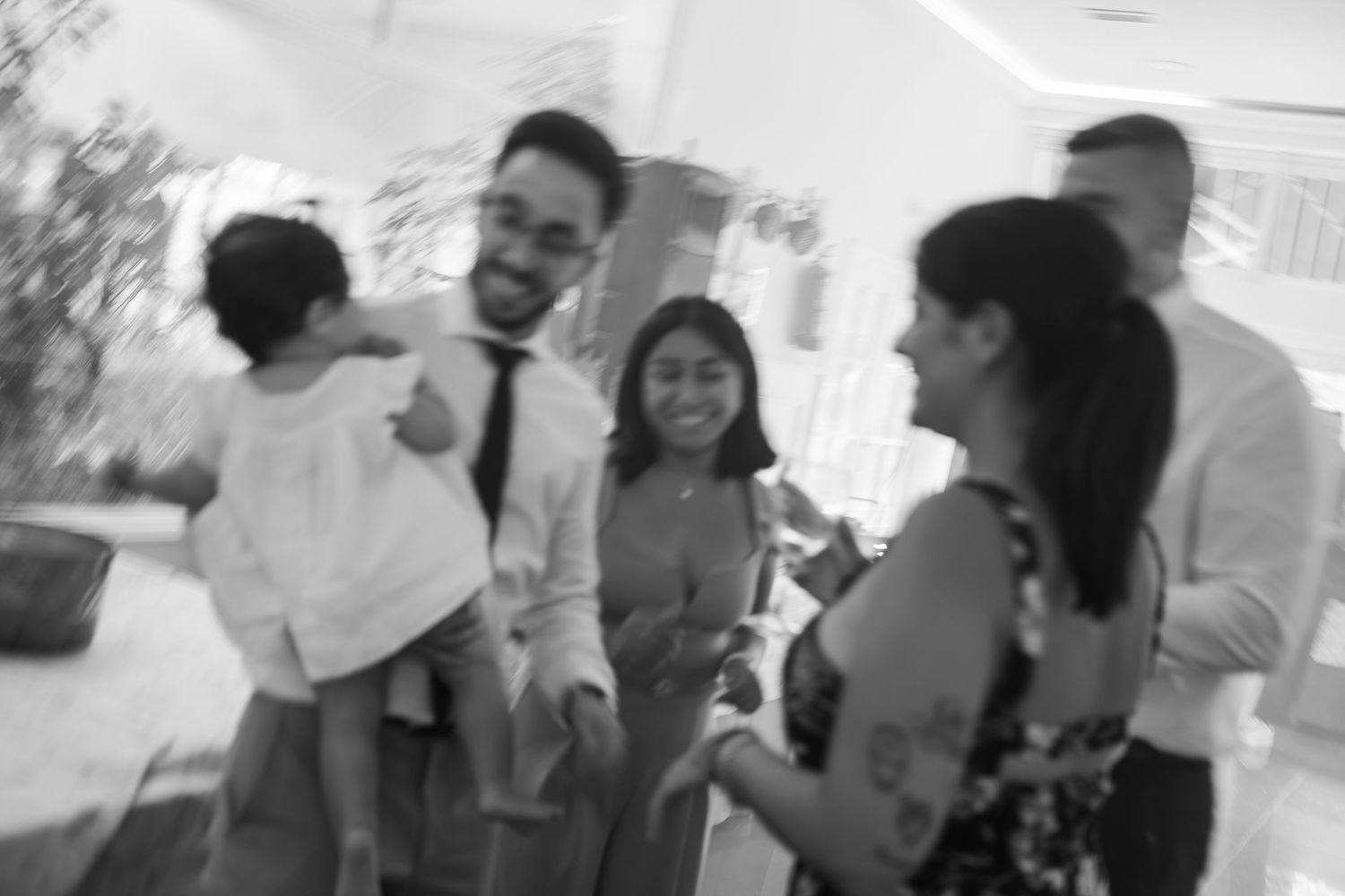 Black and white photo of a smiling group of adults chatting at a gathering while a man in glasses holds a toddler in a white dress.
