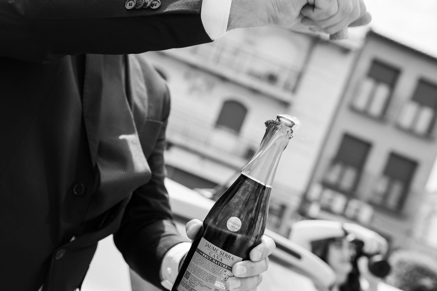 Black and white photo of a man in a suit opening a champagne bottle outdoors with city buildings in the background.