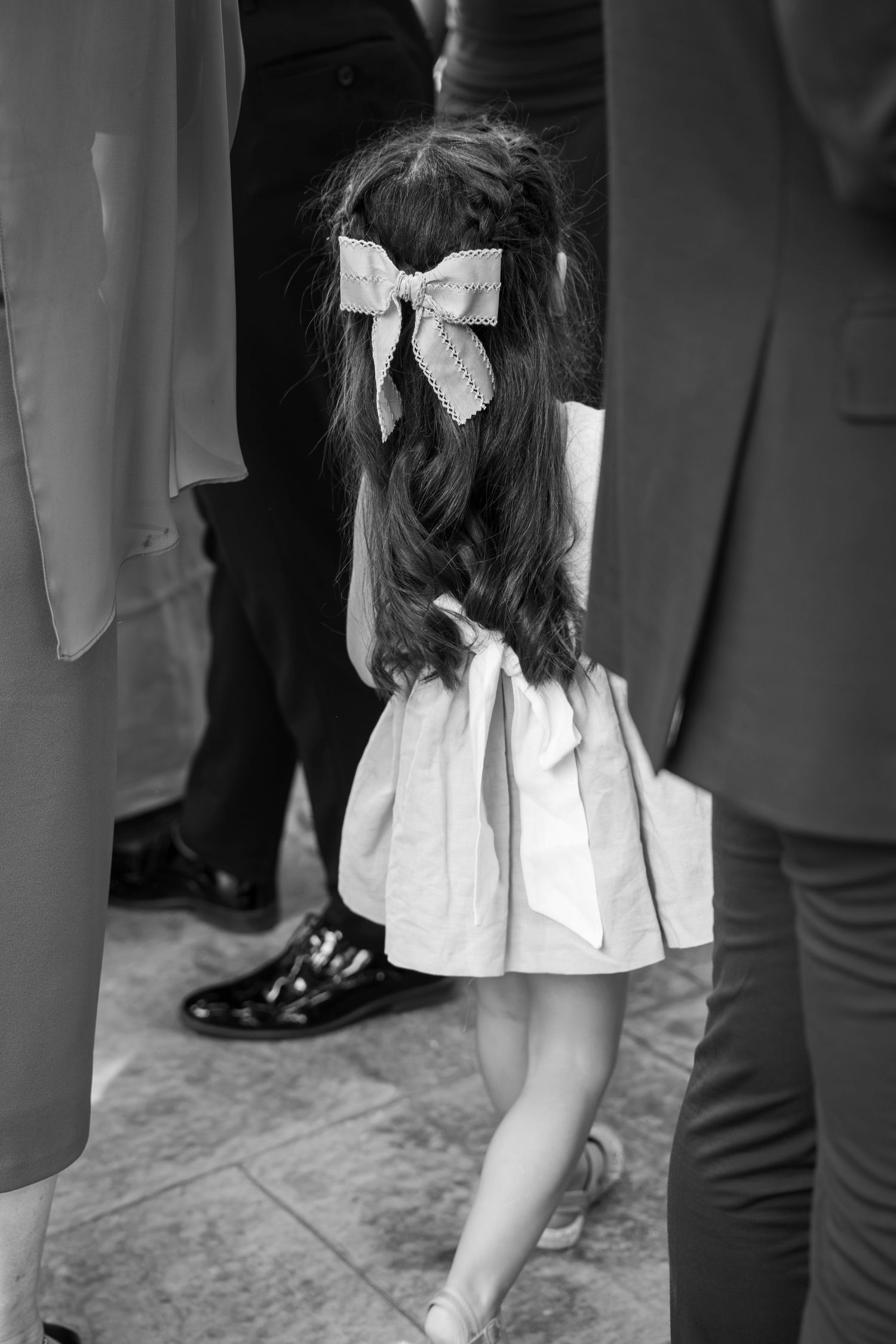 Little girl with long wavy hair and a large bow walks away in a bow-tied dress among formally dressed adults in a black and white photo.