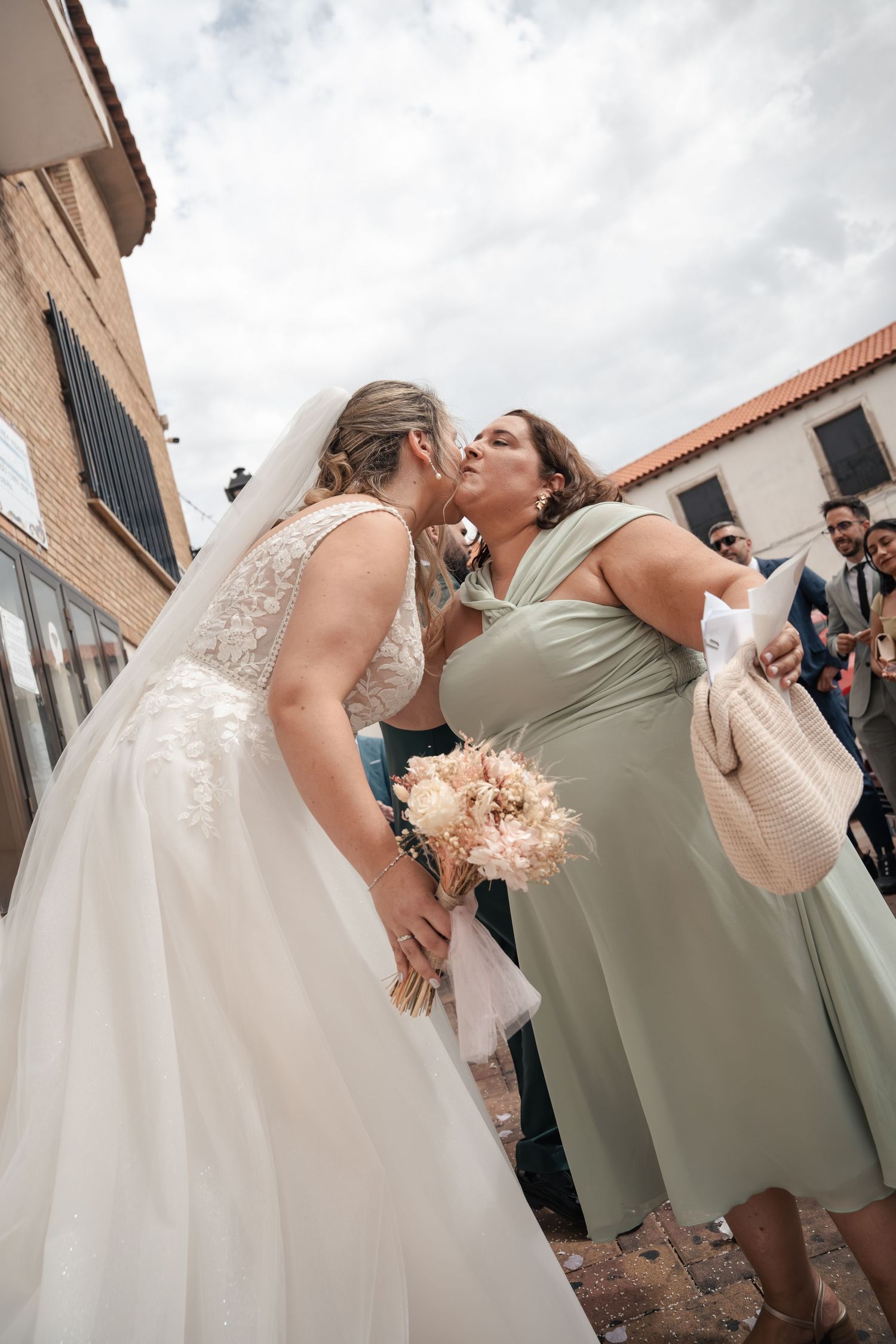 A bride in a lace wedding gown and veil kisses a guest in a sage green dress outdoors, holding a pastel bouquet, with guests nearby.