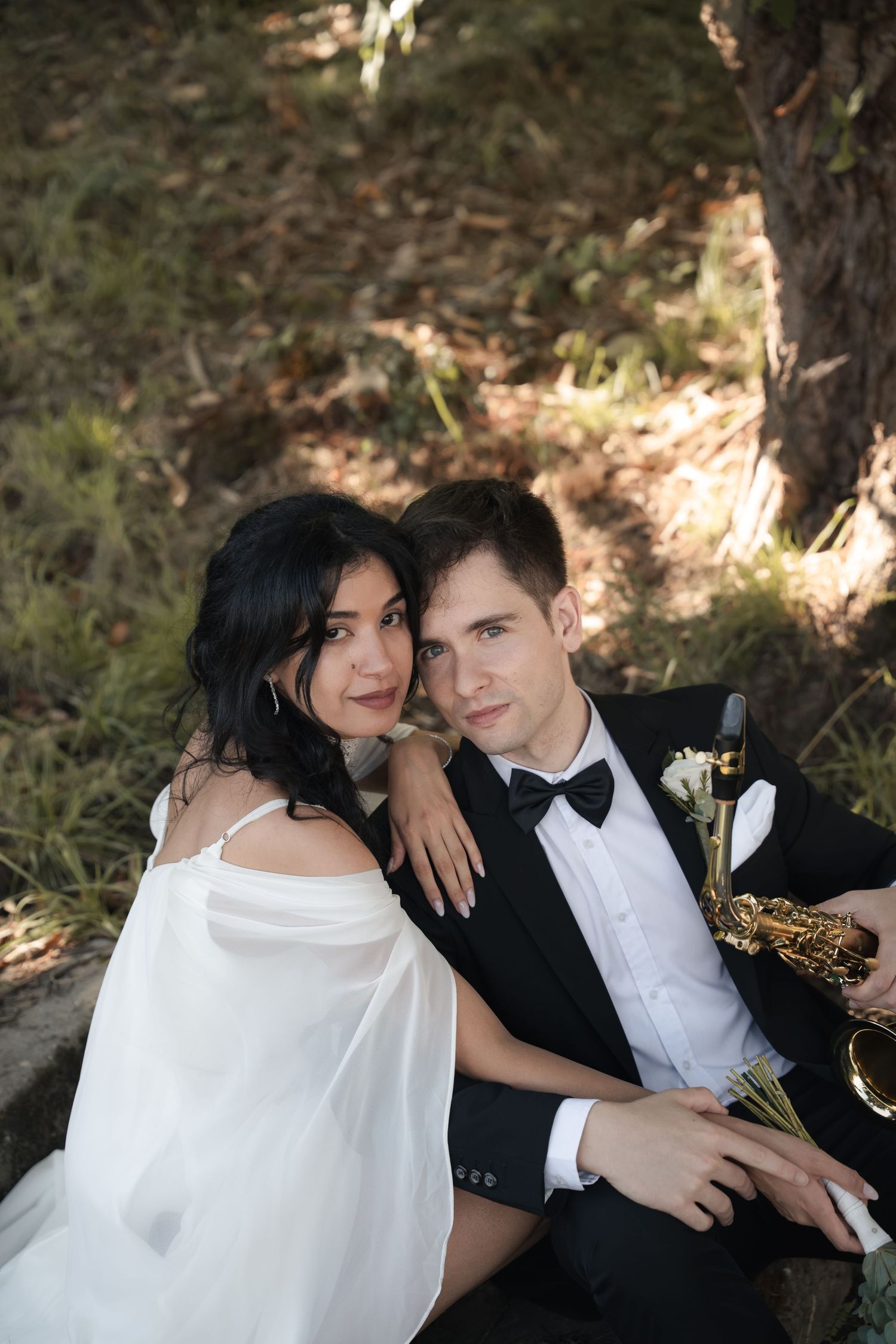 Couple in wedding attire posing close together outdoors, the groom in a black tuxedo and bow tie holding a saxophone beside the bride.