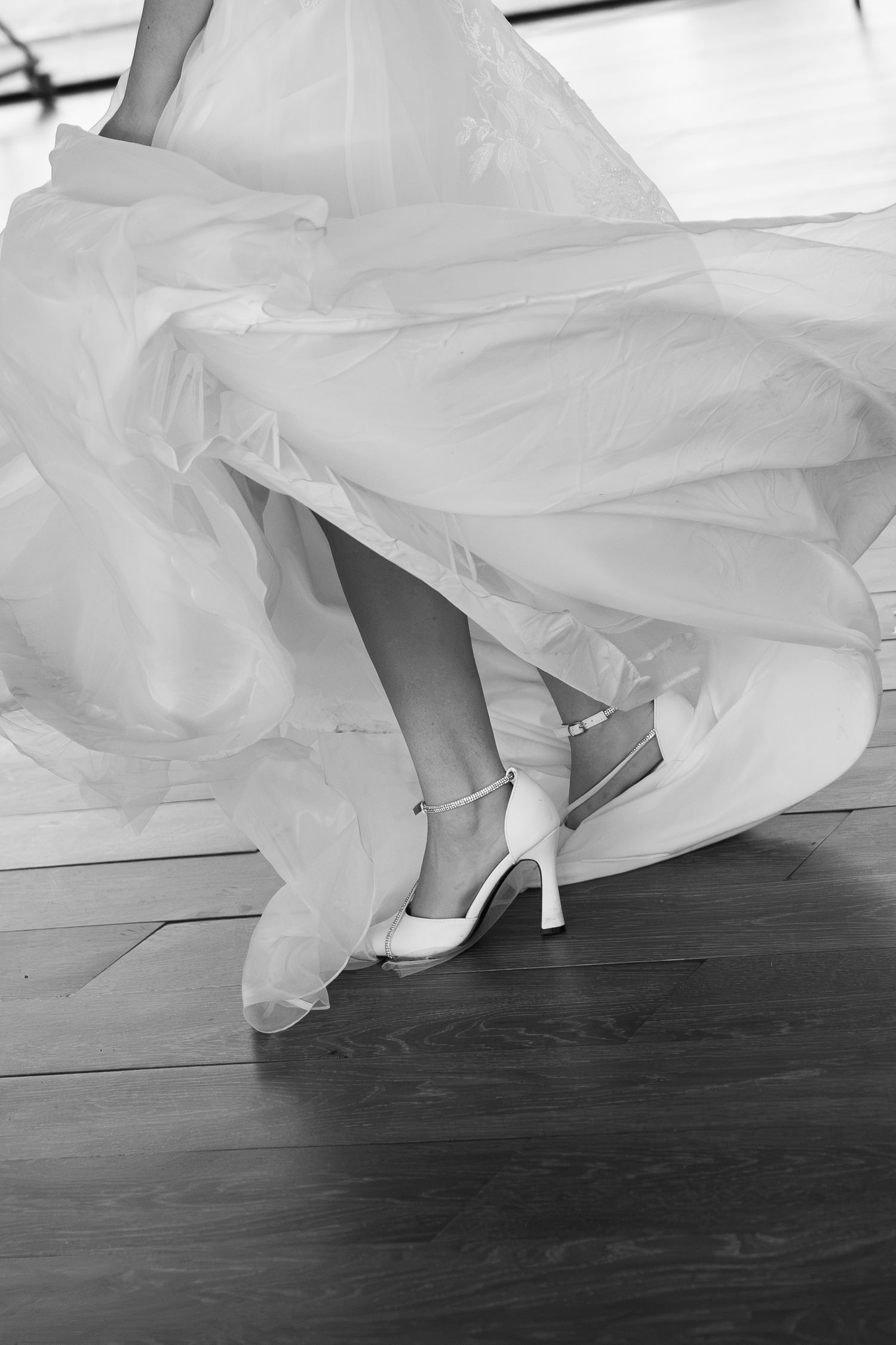 Black and white photo of a bride's flowing tulle wedding gown swirling over wooden floors, showing white ankle-strap bridal heels.