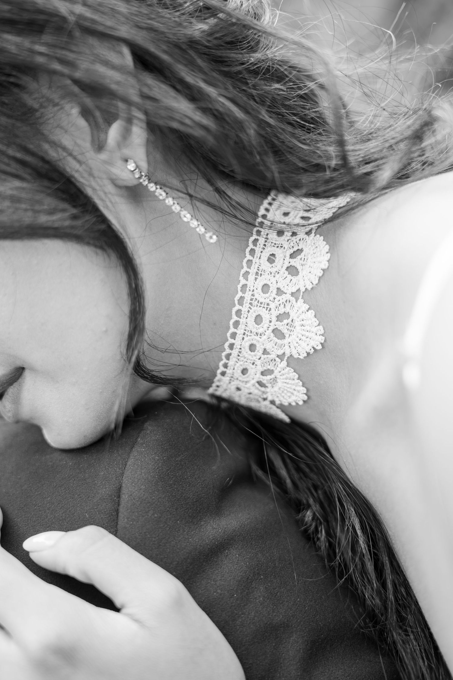 Black and white photo of a bride with lace dress detail, diamond earring, and braided hair resting on groom's shoulder.