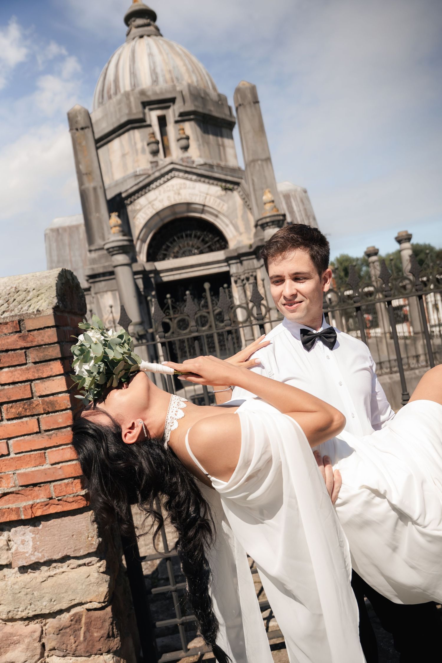 Groom in white suit carries bride in wedding dress outside historic stone church on sunny day.