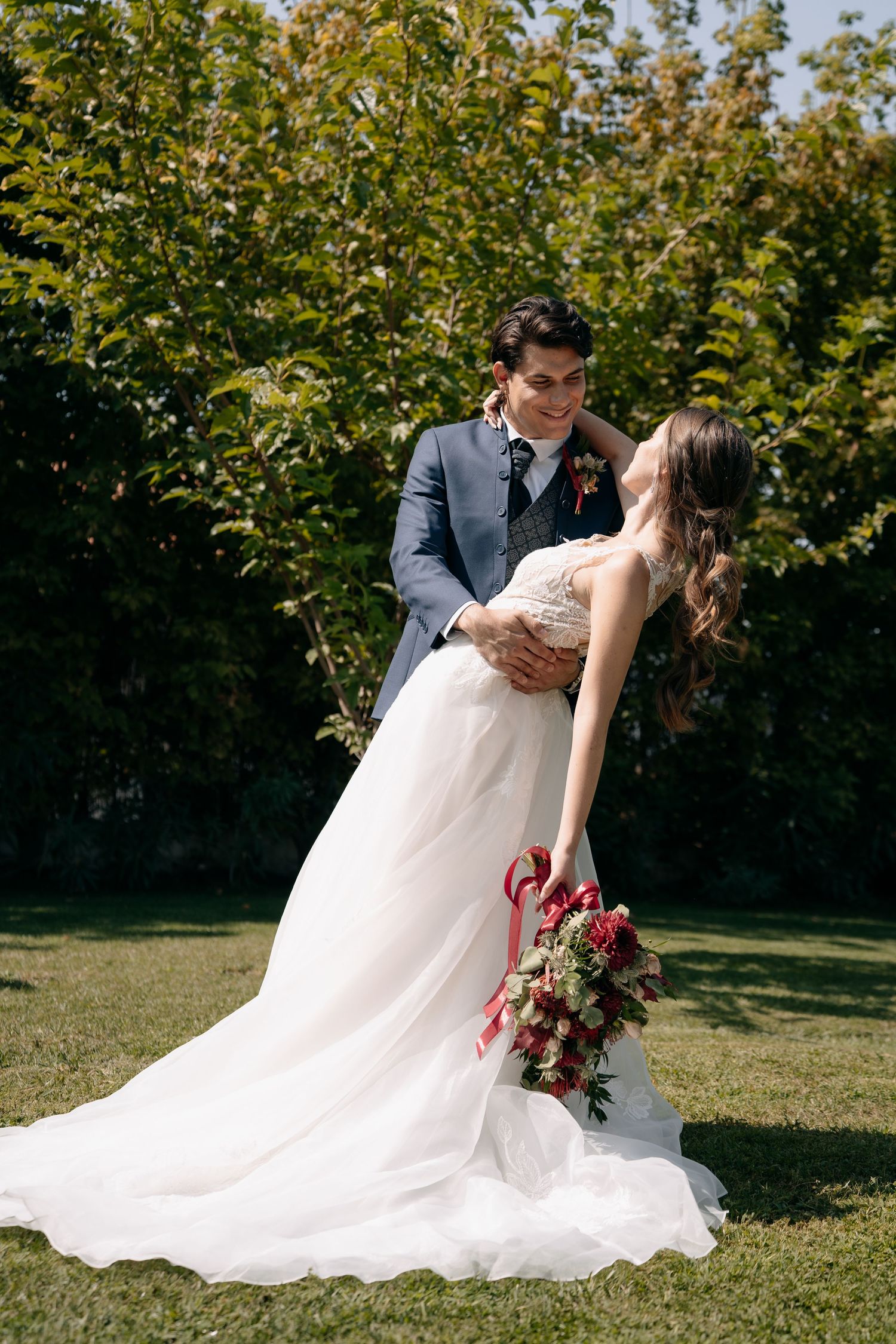 A bride in a white gown holds a bouquet while embracing her groom in a blue suit outdoors in a lush green garden.