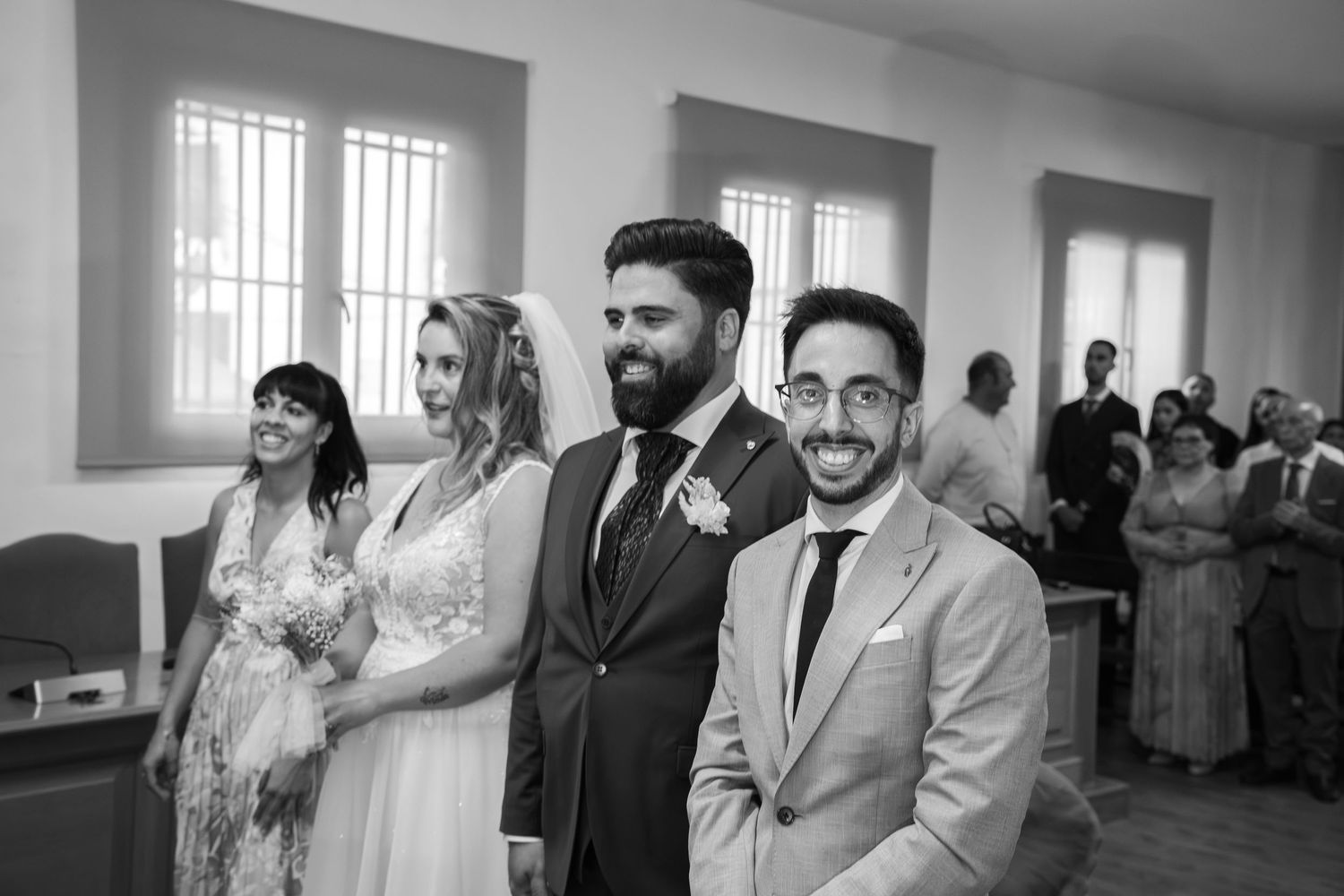 Smiling bride, groom and two guests pose together in a bright room during a wedding ceremony, with onlookers gathered in the background.