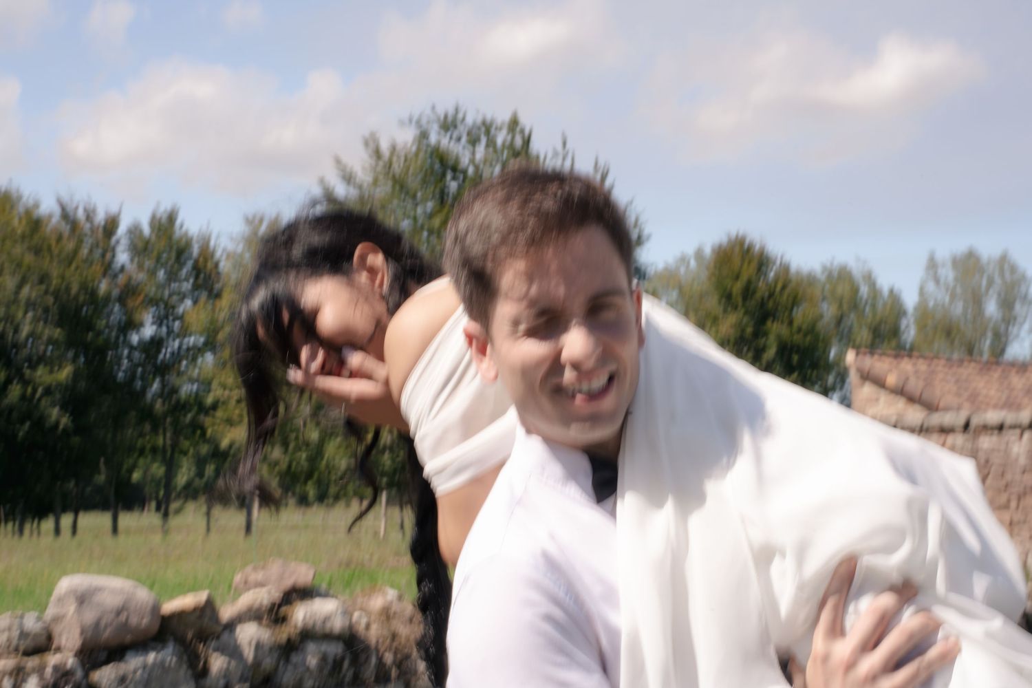 A smiling man in a white shirt carries a dark-haired woman over his shoulder in a sunny grassy field with trees and a stone wall.
