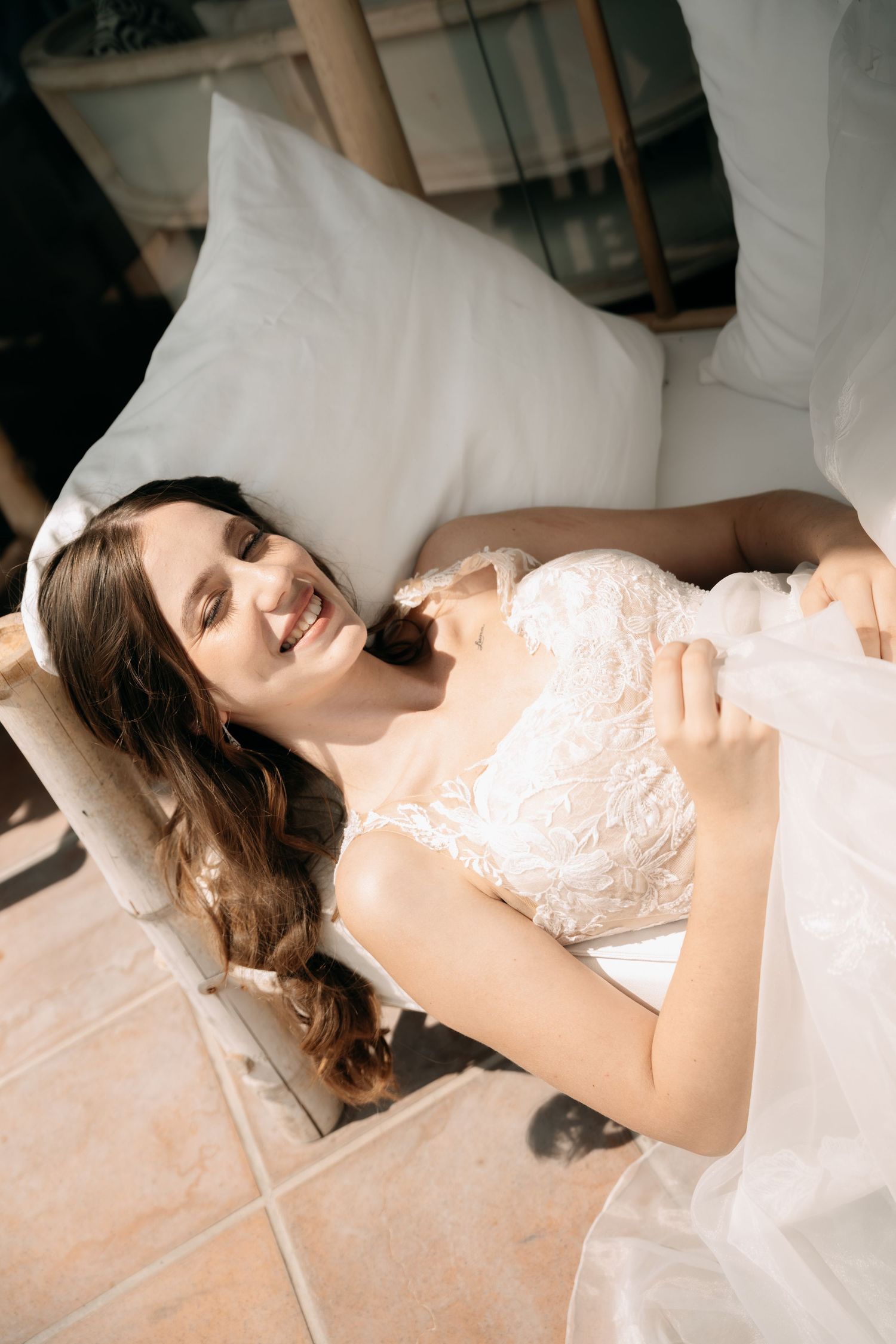 Smiling bride in a lace wedding gown with long wavy hair relaxes on a cushioned outdoor lounger in warm sunlight.