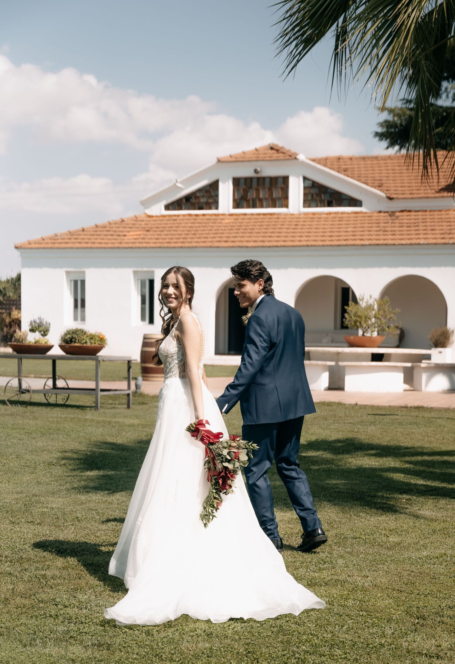 Bride in a flowing white gown holding a red bouquet walks hand in hand with a groom in a navy suit on a lawn near a white villa.