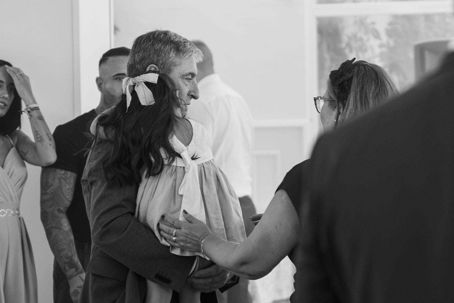Black and white photo of a man in a suit hugging a young girl in a white dress with a bow at a wedding while guests embrace nearby.