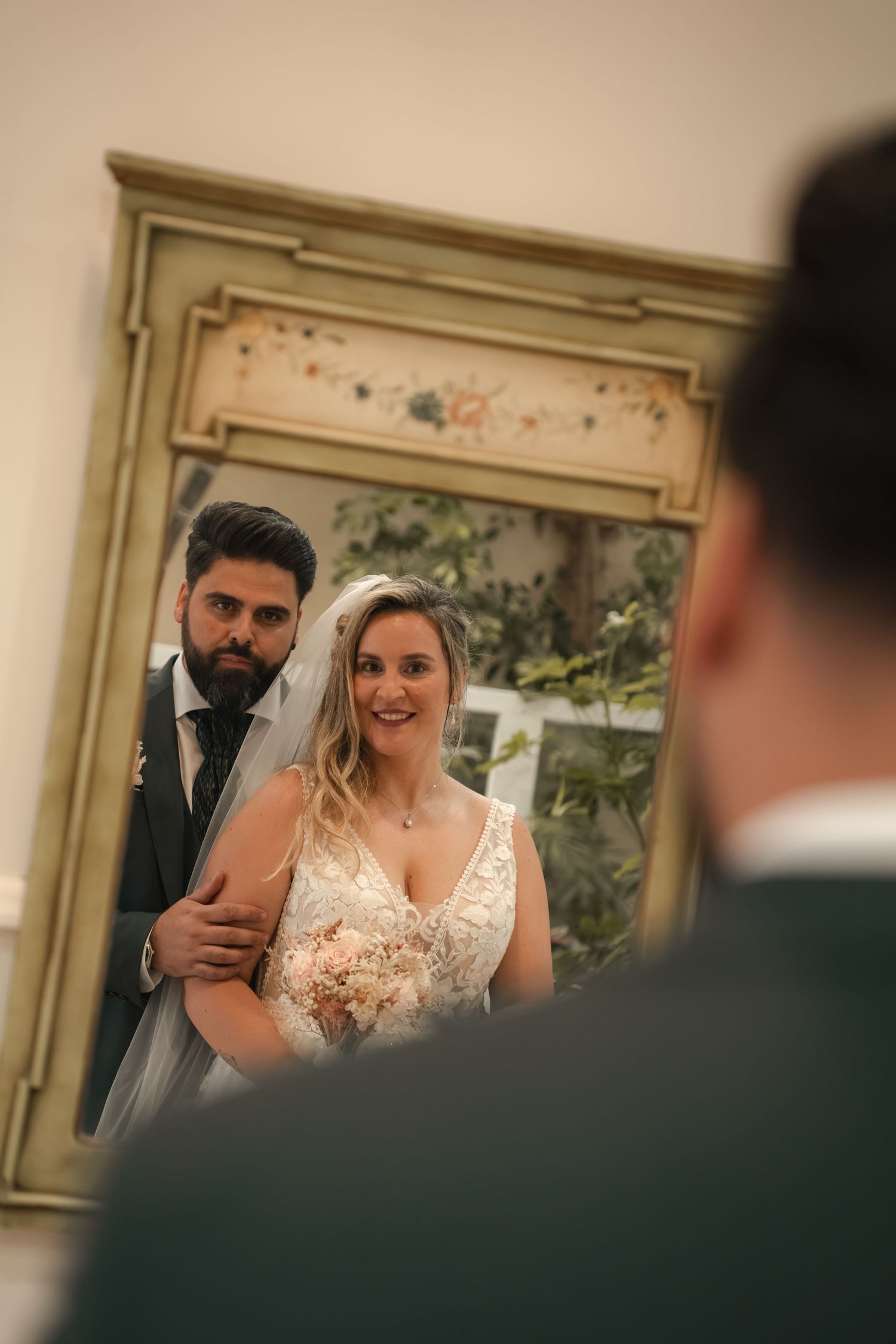 Smiling bride in a lace gown and veil holding a bouquet, embraced by her groom, reflected in an ornate vintage mirror at their wedding.