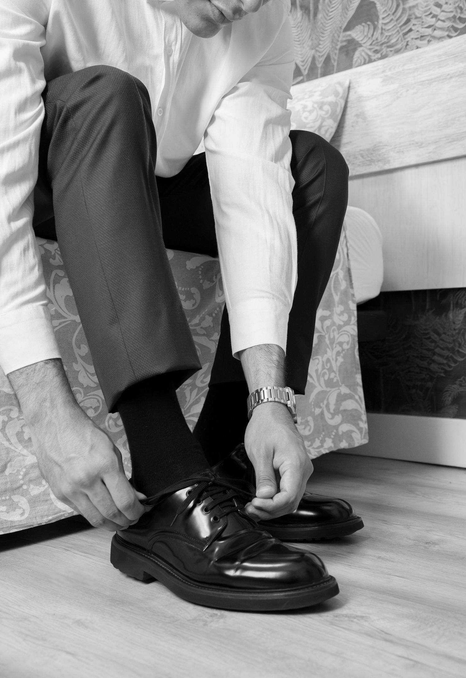 Black and white photo of a man in a dress shirt and trousers sitting on a bed, tying his polished leather shoe.