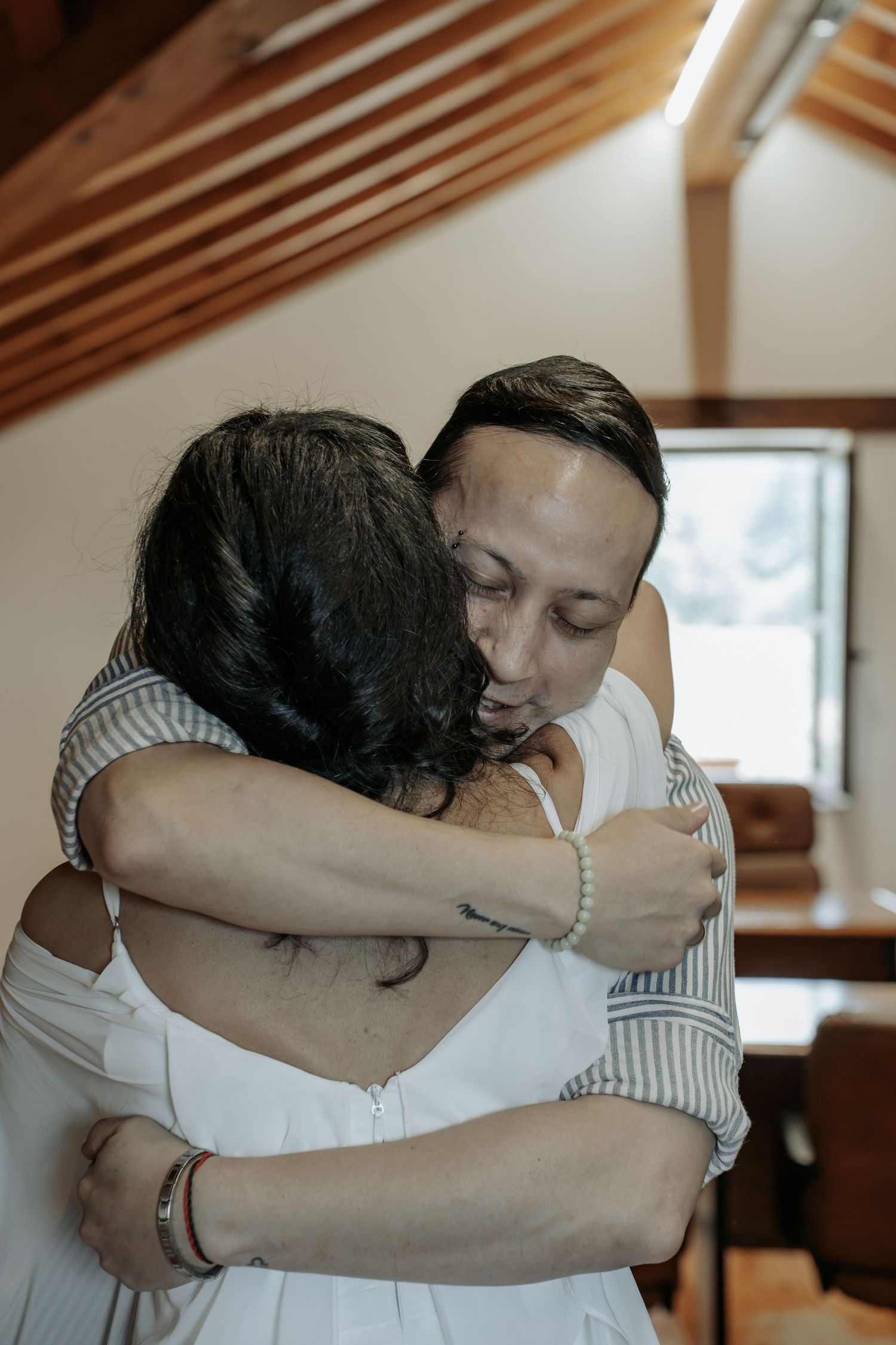 Man with closed eyes embraces a woman in a white dress in a warm, sunlit room with wooden beams.
