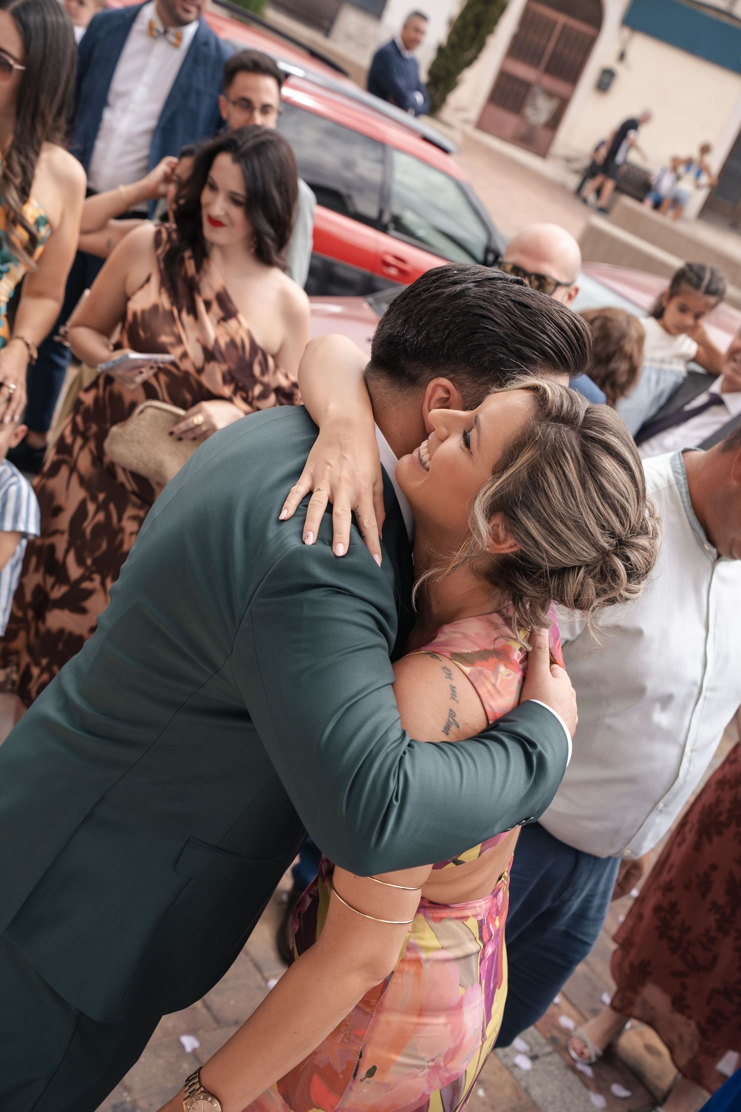 A man in a green suit hugs a smiling woman in a colorful dress at an outdoor gathering while guests in elegant attire look on.