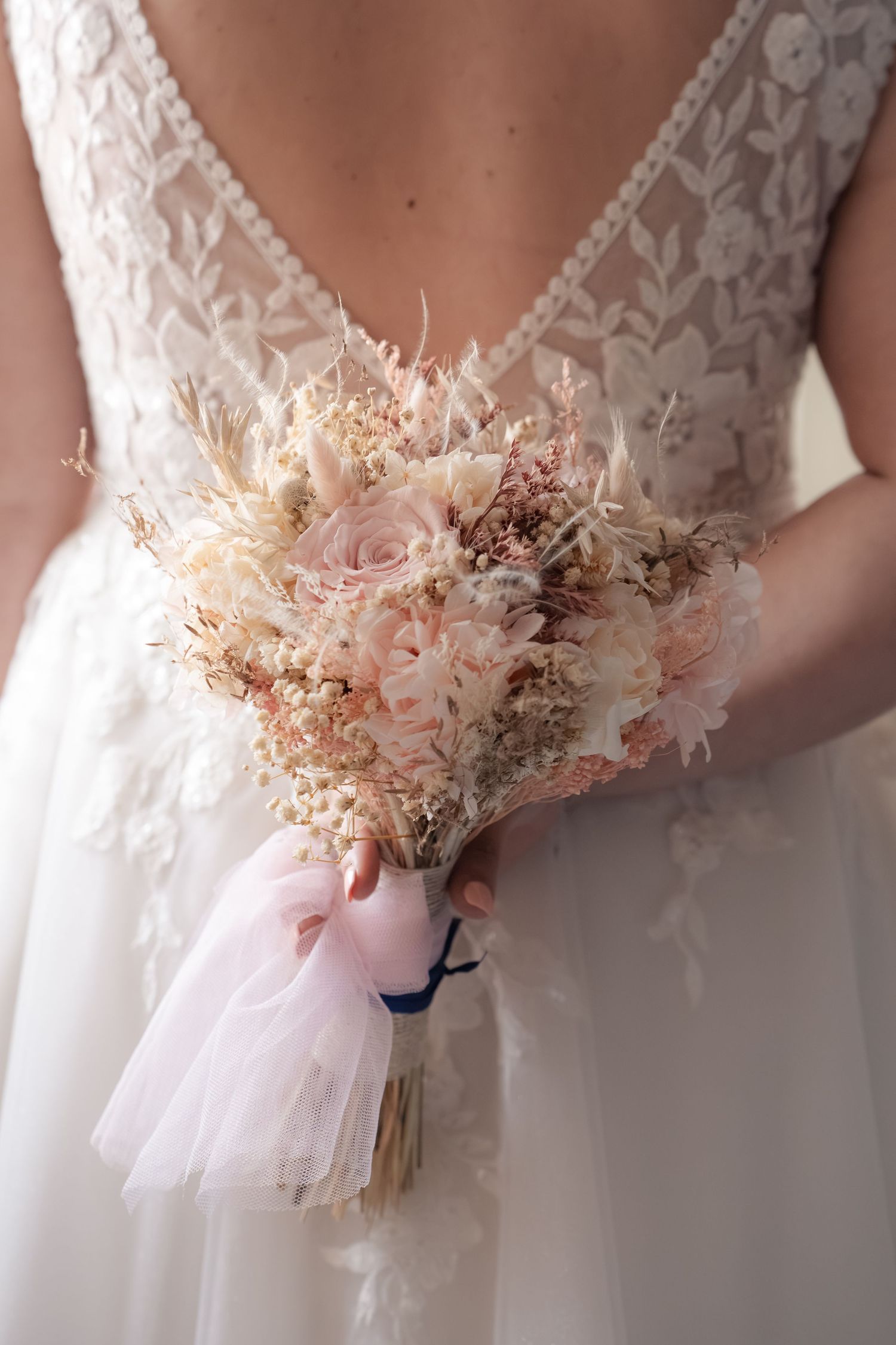 Bride in a lace wedding gown with a low back holds a blush pink and dried flower bouquet tied with a tulle ribbon.