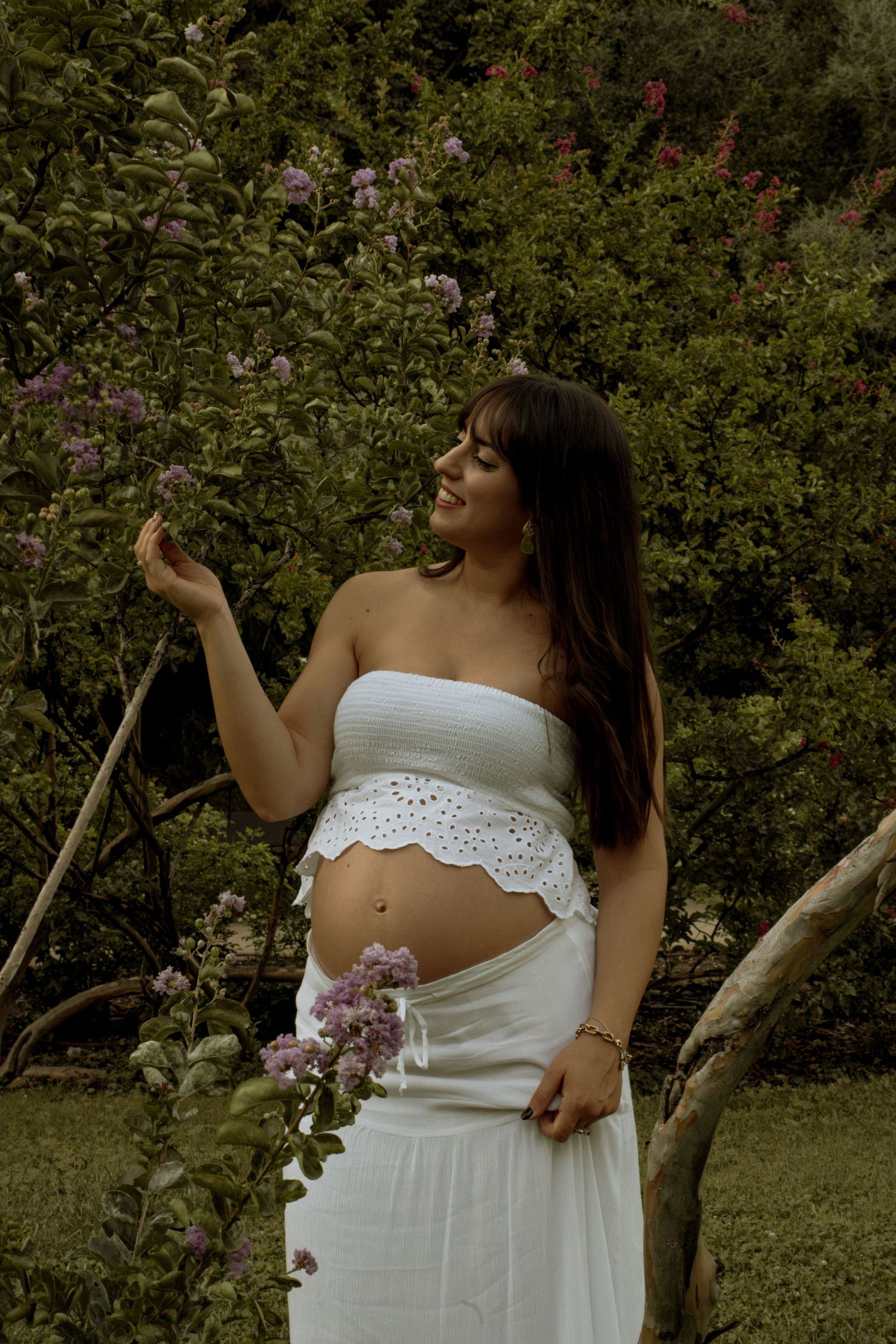 Pregnant woman in white eyelet crop top admires purple flowers while standing near rustic wooden branches.