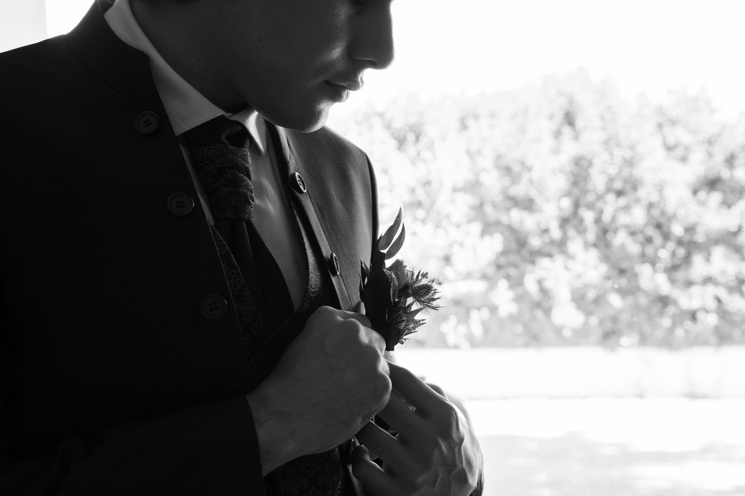 Black and white photo of a groom in a suit and tie adjusting his boutonniere on his lapel, with soft sunlit trees in the background.