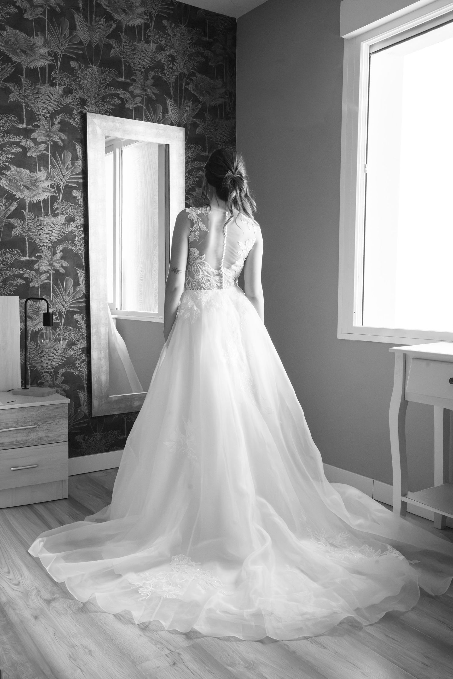 Black and white photo of a bride in a lace-back wedding gown with a long flowing train, facing a mirror beside a bright window.
