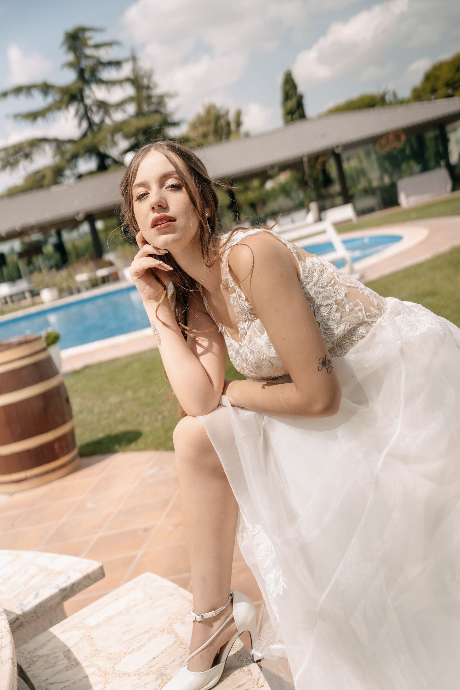 Bride in a lace and tulle wedding dress with white ankle-strap heels sits on stone steps beside a pool and garden.