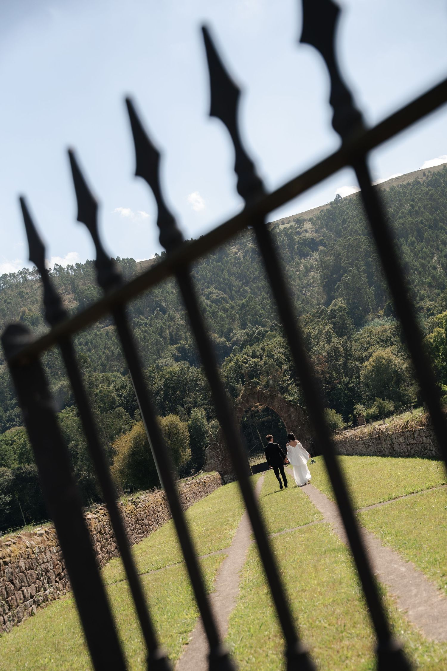 Bride and groom walk hand in hand along a grass path toward a stone arch, framed by an ornate black iron gate and forested hills.