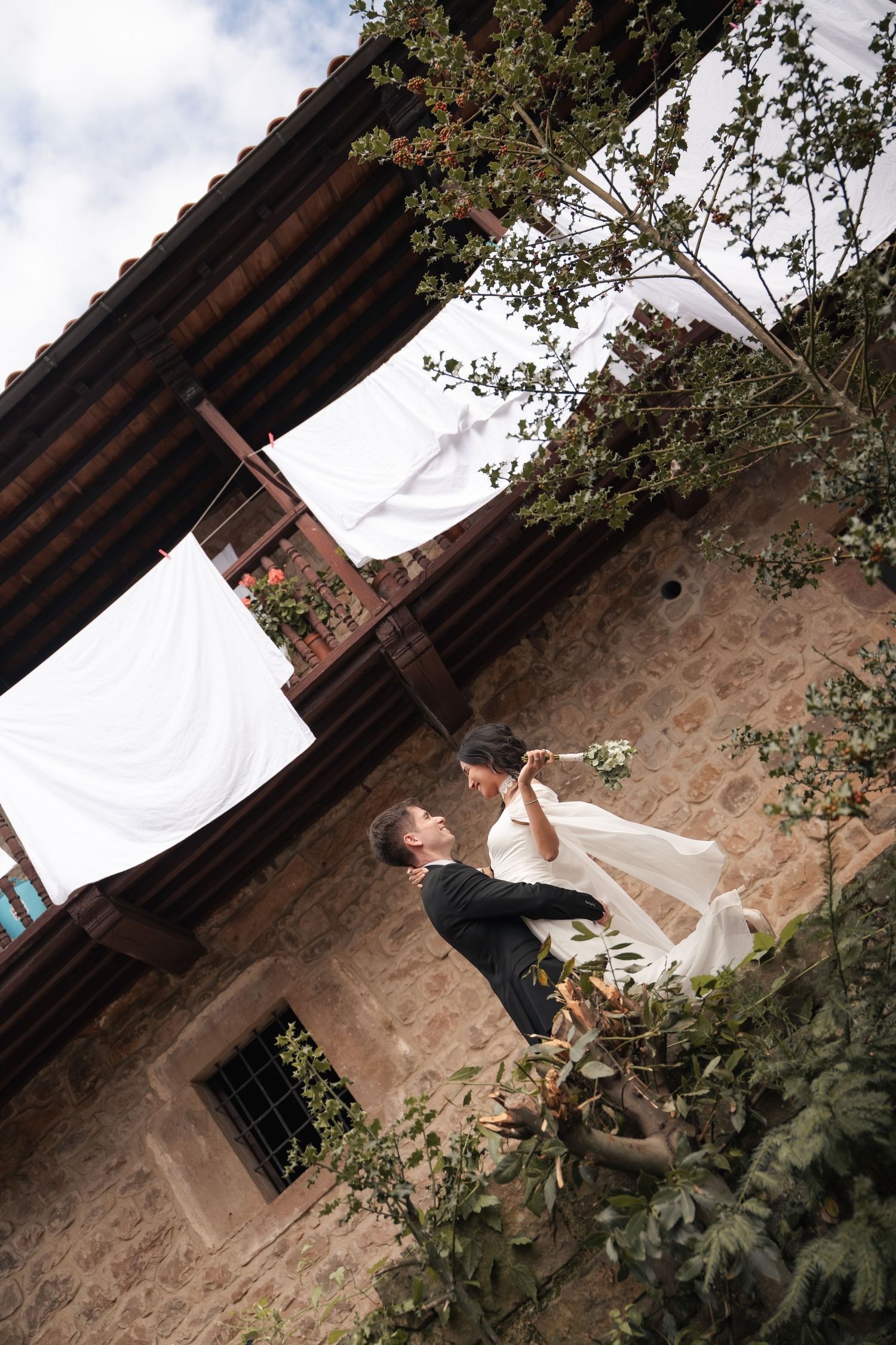 Bride and groom share a romantic moment outdoors in front of a rustic stone building with wooden balconies and white linens hanging above.