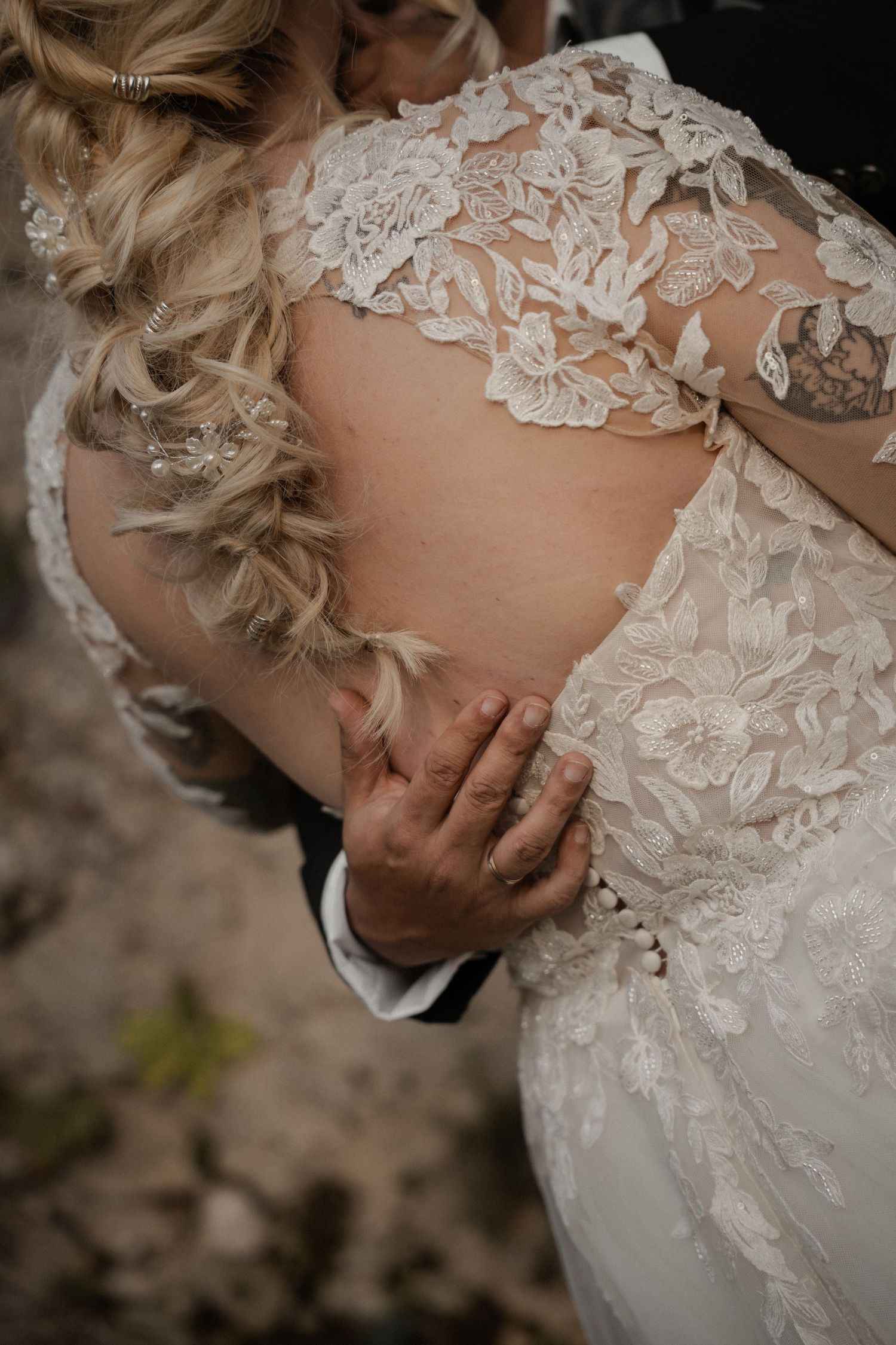 Close-up of groom's hand on bride's lace wedding dress back, showcasing intricate floral lace detailing and pearl buttons.