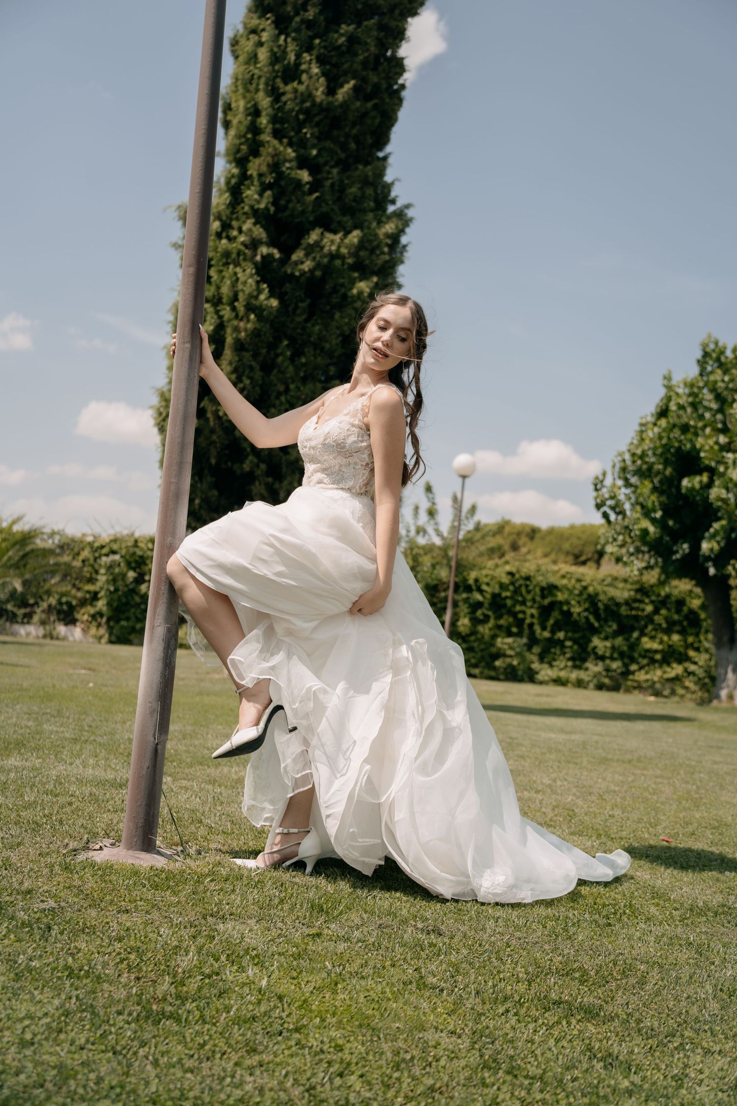Bride in a flowing white tulle wedding gown with a lace bodice leans on a metal pole on a sunny green lawn, one heeled foot raised.