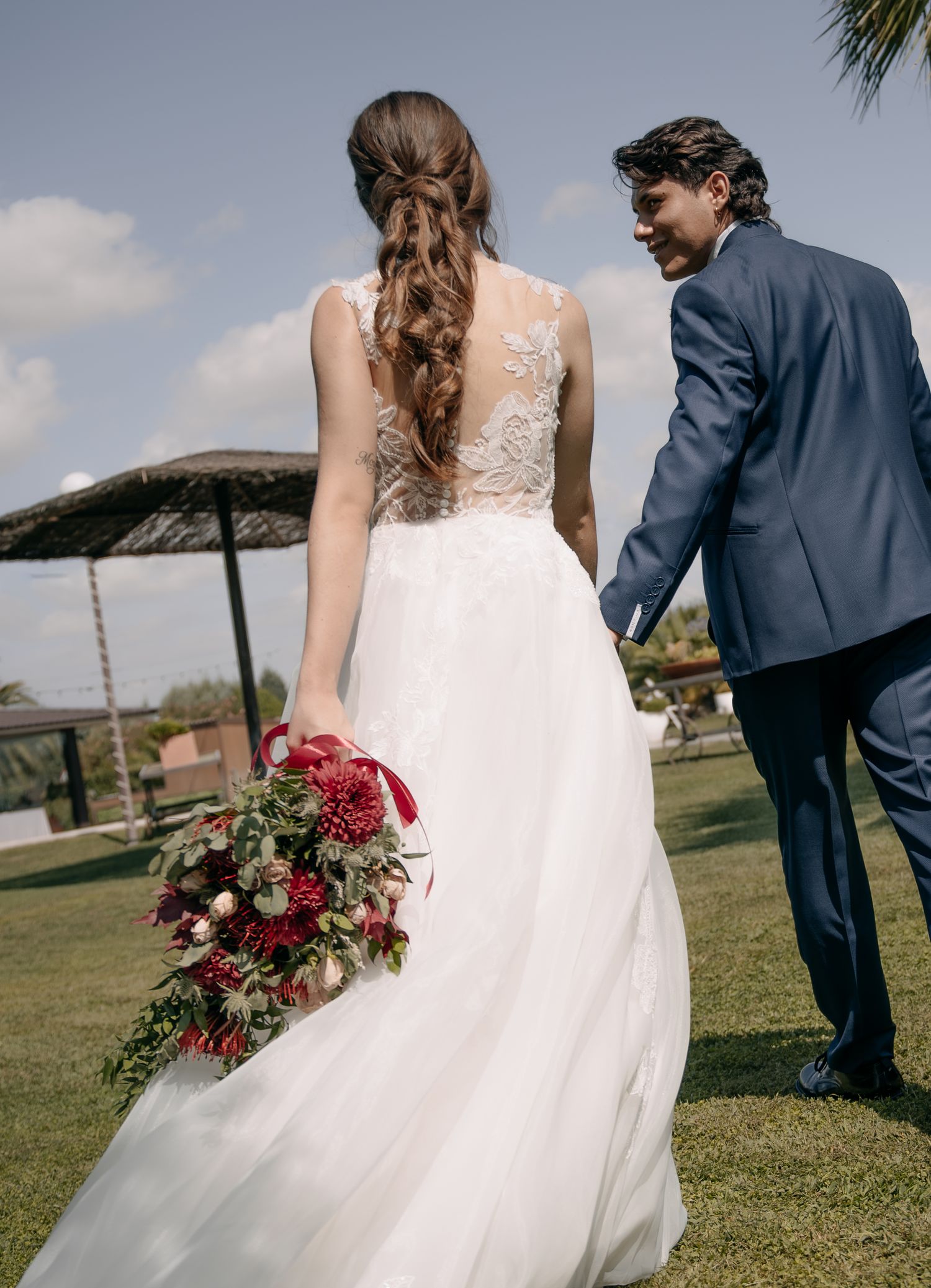 Bride in a lace-back wedding gown with a braided updo holds a red floral bouquet while walking hand in hand with a groom in a blue suit.