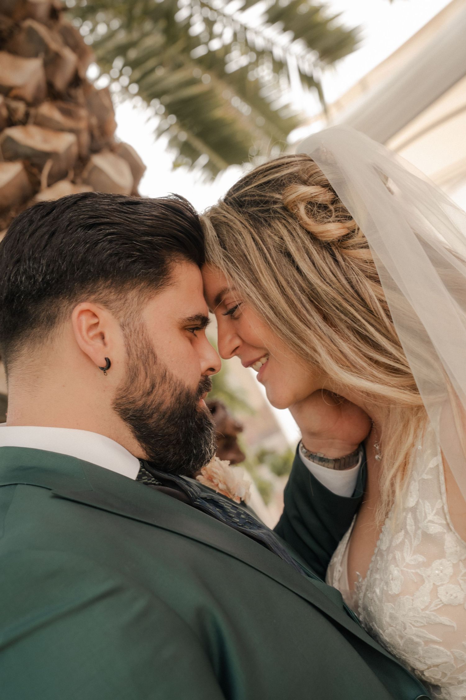 Smiling bride in a lace gown and veil touches foreheads with her bearded groom in a green suit beside a palm tree.