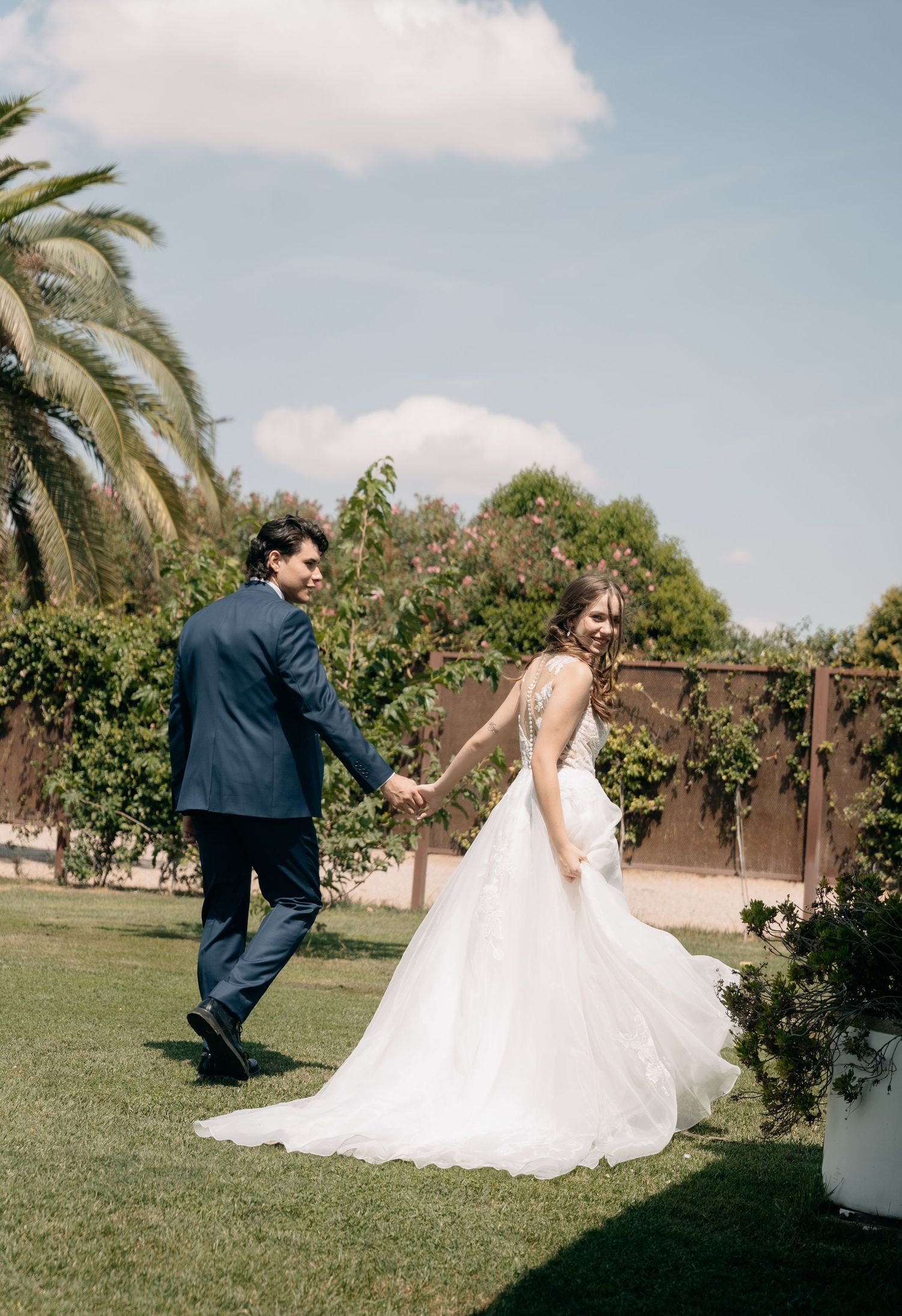 Bride in a flowing white gown and groom in a navy suit hold hands and walk across a sunny garden lawn on their wedding day.