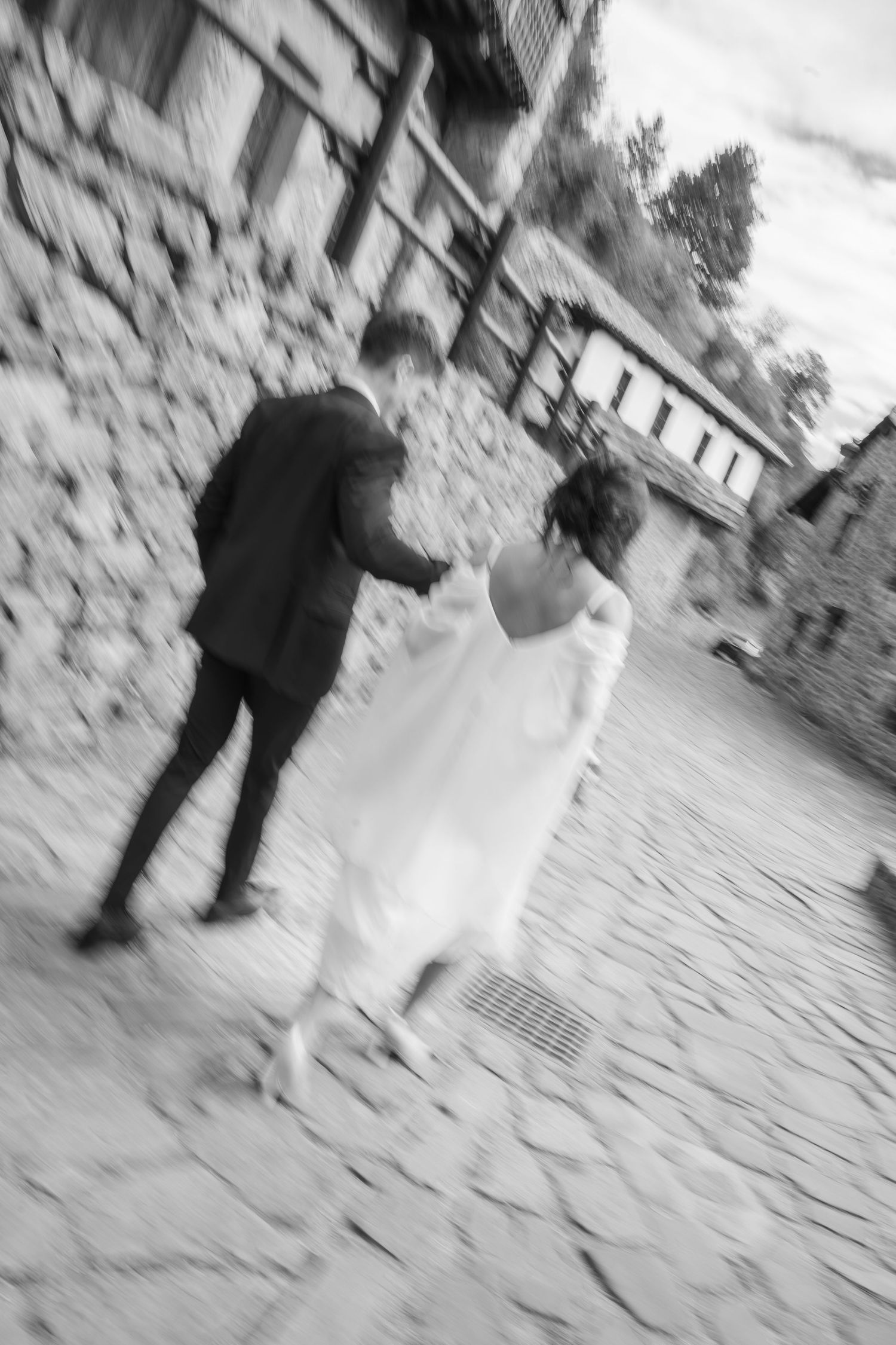 Black and white photo of a bride and groom walking away on a cobblestone street beside old stone buildings, captured with motion blur.