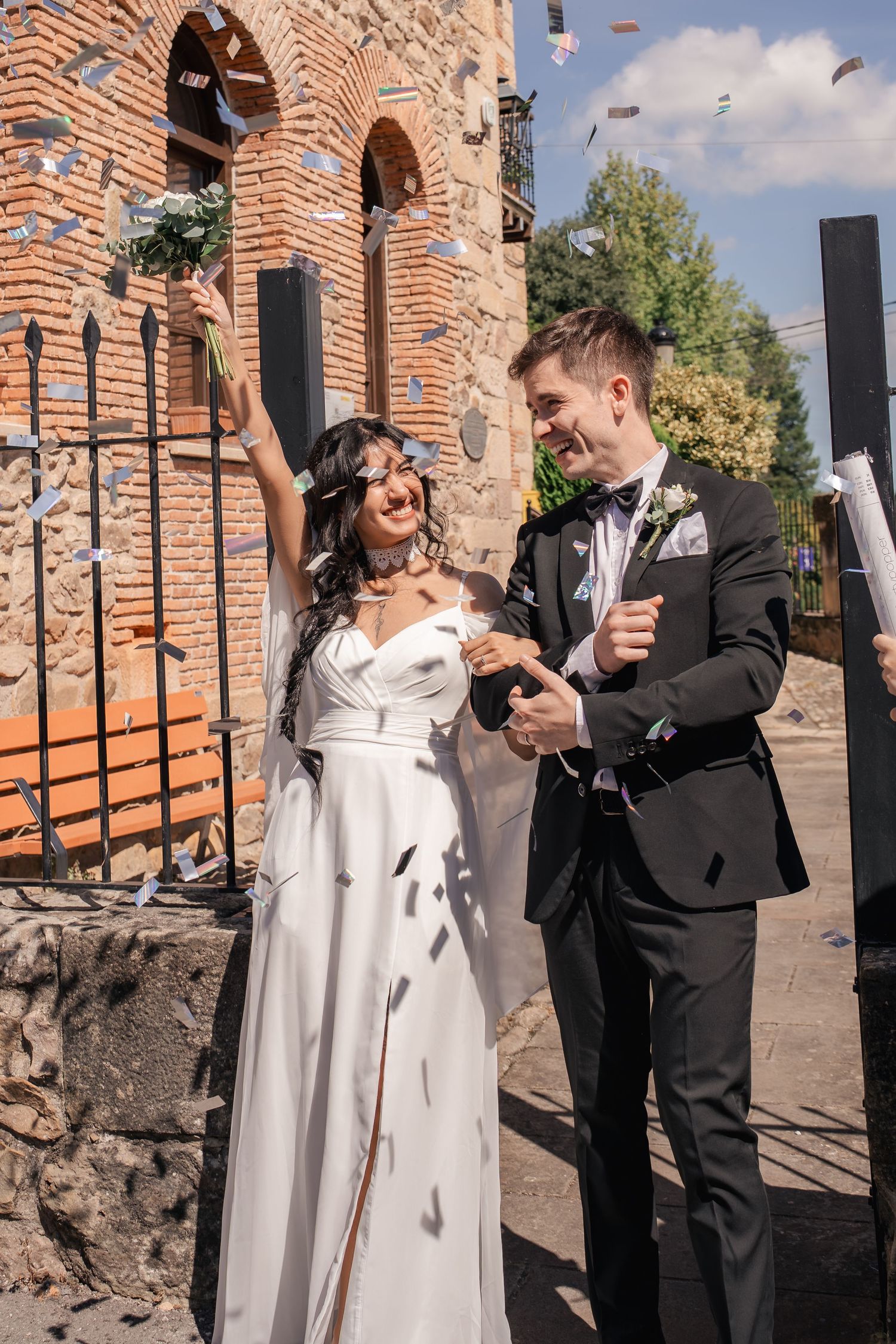 Smiling bride in a white gown and groom in a black suit walk outside a brick building as guests toss confetti on their wedding day.
