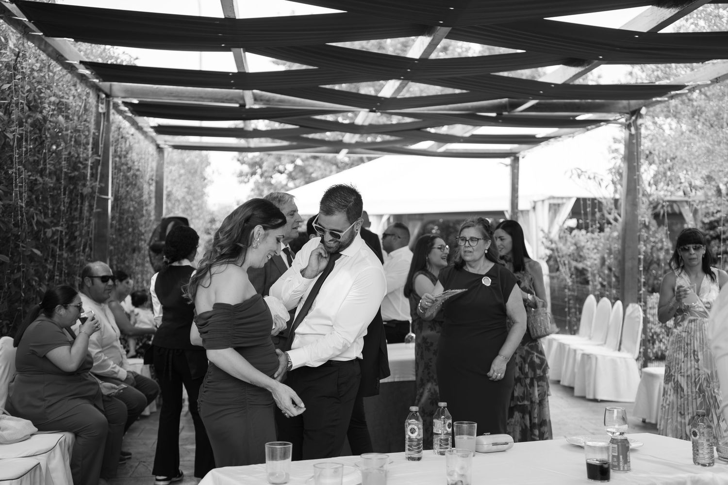 Black and white photo of wedding guests mingling and chatting at an outdoor reception with pergola shade and set tables.