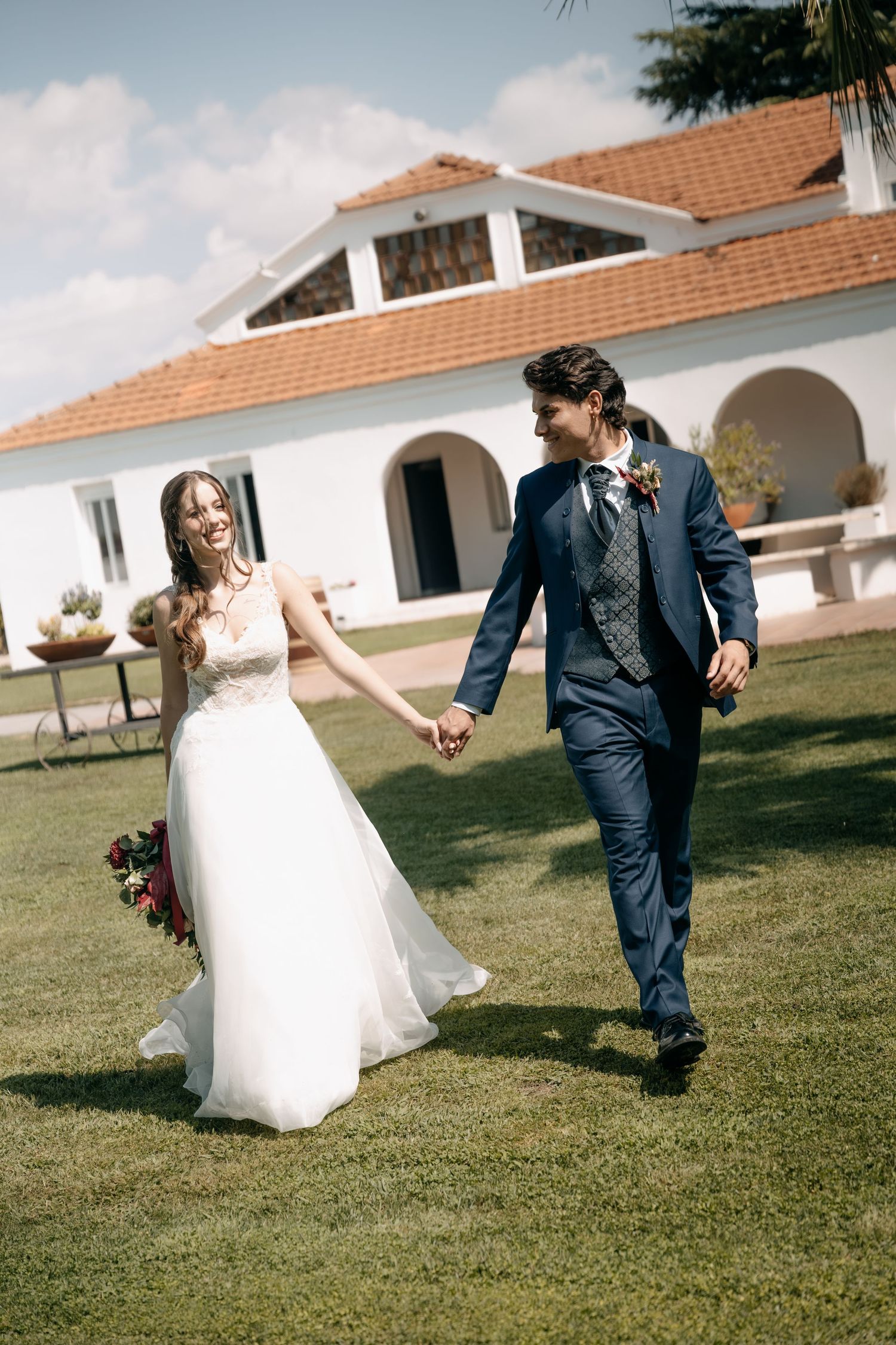 Smiling bride in a white lace gown and groom in a navy suit hold hands and walk across a sunny lawn near a Mediterranean-style villa.